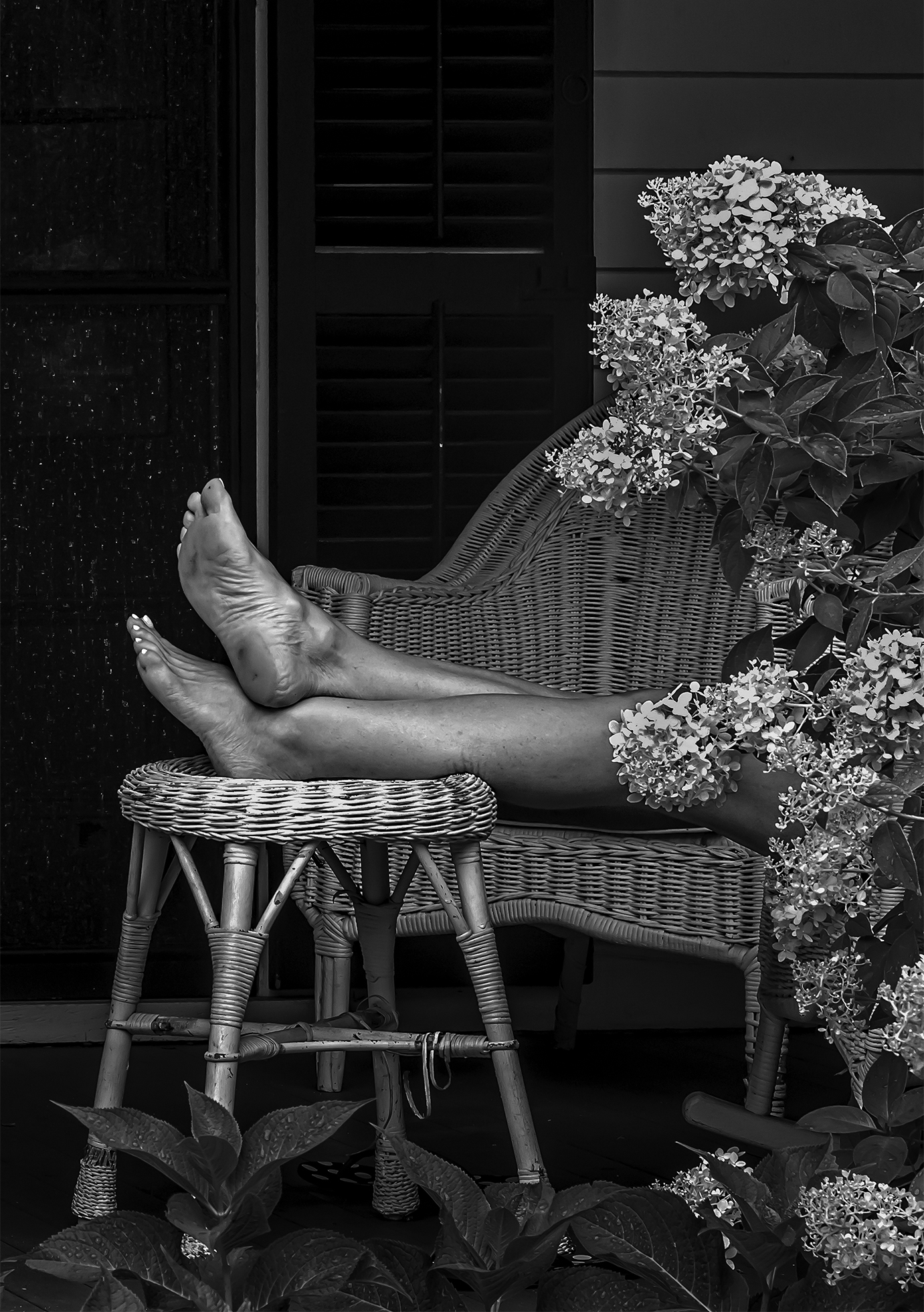 A woman sits on a wicker loveseat on a patio with her bare legs extended and crossed, as her feet rest atop a small, round wicker table. A flowering bush with large, cone-shaped clusters of delicate flowers cascade in front of the woman on the right.