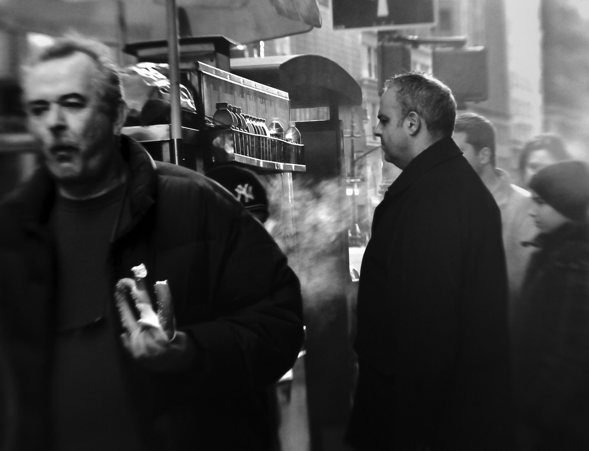 A line of men wait at a pretzel cart in a city. A man walks away from the cart toward the camera with a soft pretzel in his left hand. The men are in coats and there is steam in the air.