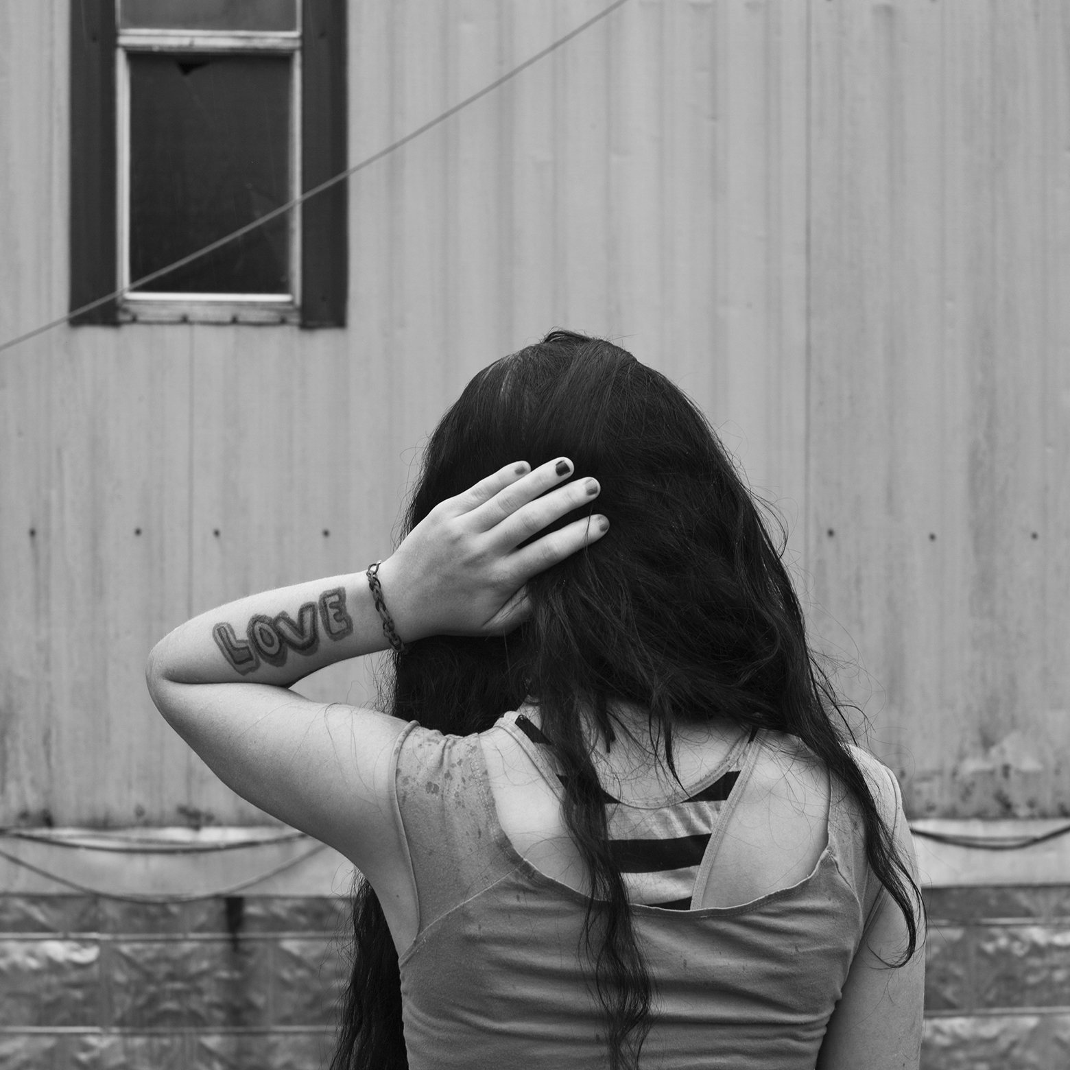 The top half of a young woman with long, dark hair from the back with her left hand on the back of her head. She is wearing layered tank tops and has a prominent tattoo of the word “LOVE” on her left forearm. She is looking at a building with a window.