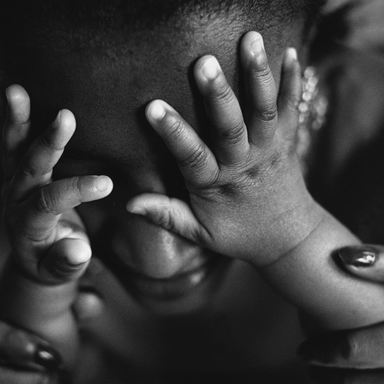 Close-up of an infant’s hands on a woman’s face obscuring her eyes. The woman is looking down on the infant and she has her hands around the infant’s forearms.
