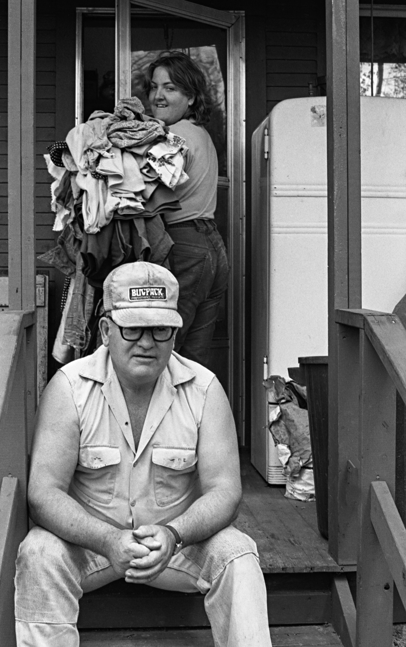 A man with a Bugpack cap and glasses sits on the top step of a porch facing the camera with his elbows resting on his knees. A woman behind him looks over her shoulder at him as she opens a door while carrying laundry. The porch has a refrigerator on it.