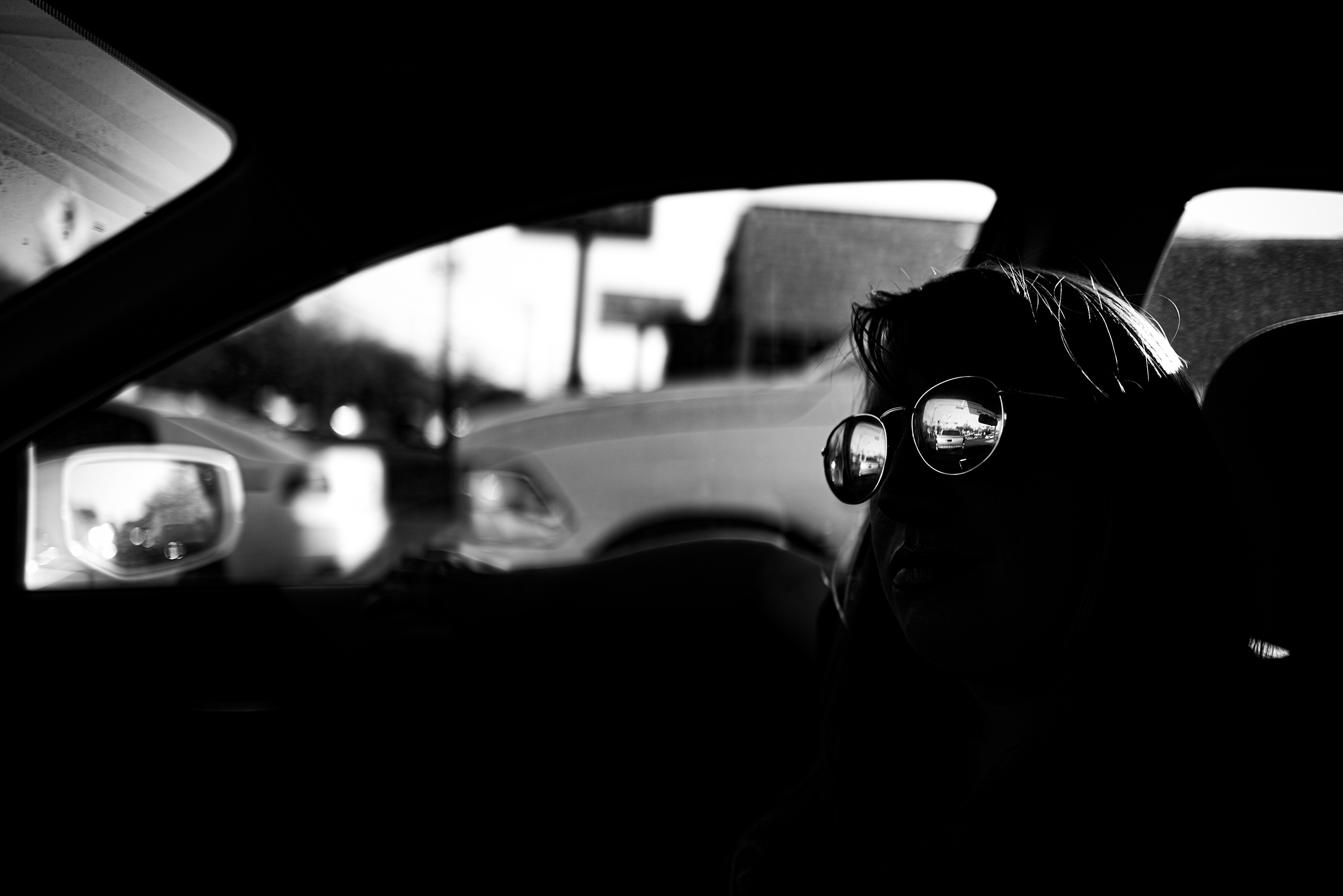 Looking into a car from the driver’s side window out to the street beyond. Everything in the car interior is black except for the shine from the sunglass lenses of a person in the front passenger seat.