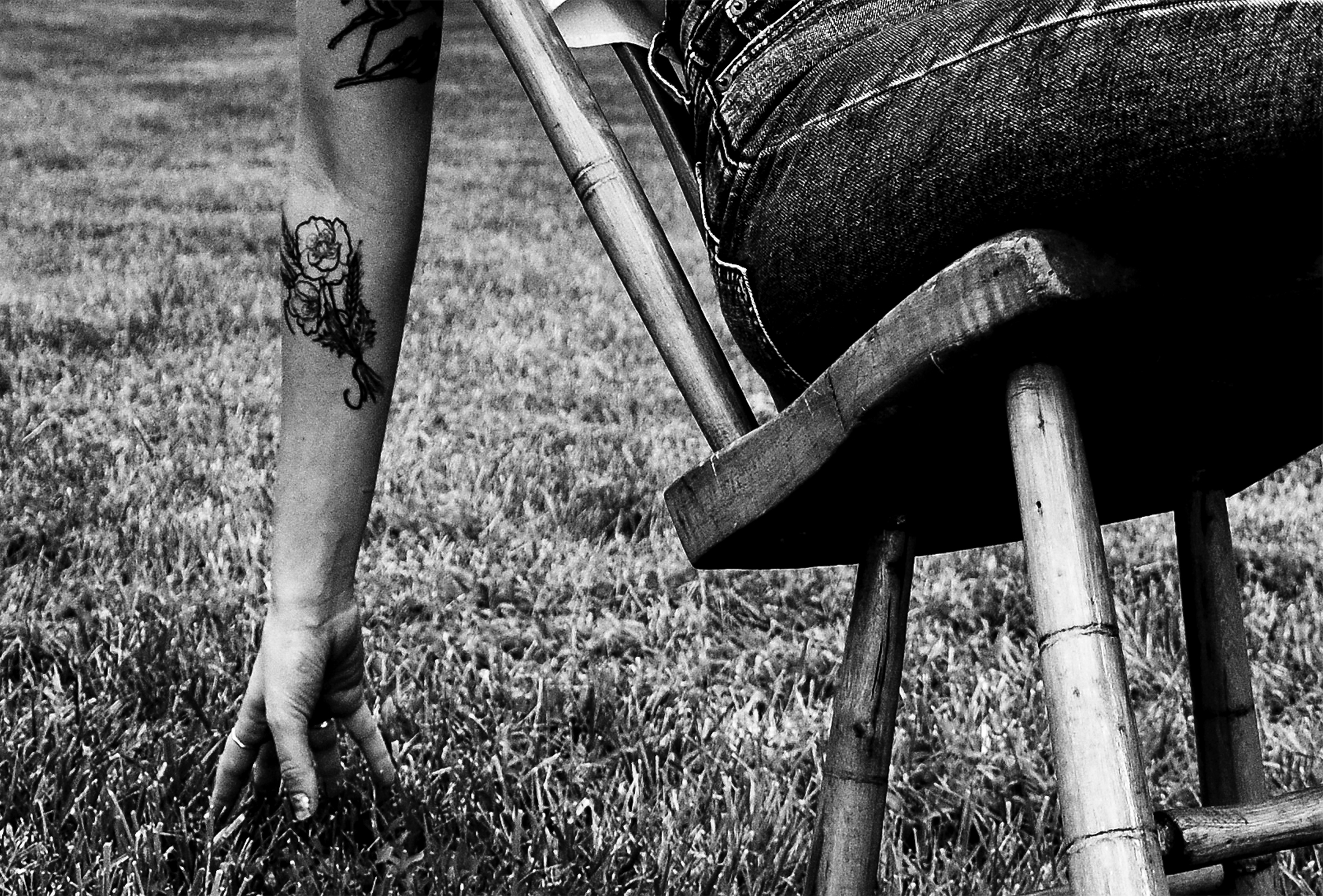 A woman in jeans with a large flower tattoo on her forearm and another tattoo on her upper arm sits on a wooden spindle-back chair in the grass and leans so far back that her fingertips are braced against the ground.