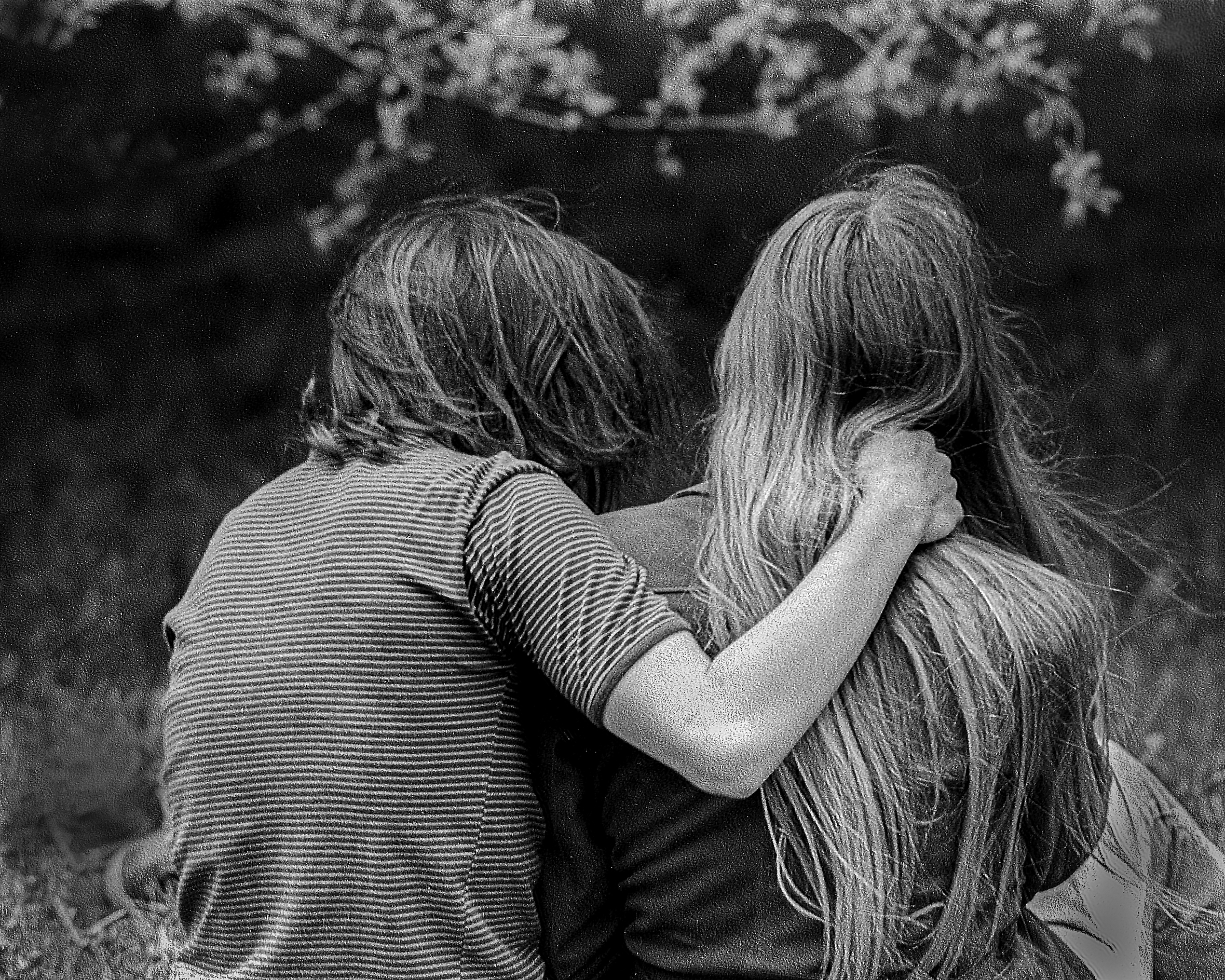 A teenage boy and girl sit next to each other on the grass, and the boy has his arm around the girl’s shoulder. They are shown from the back. He has straight, shoulder-length hair and she has straight, waist-length hair.
