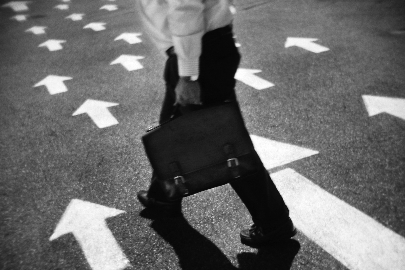 A man shown from the neck down in business attire carrying a briefcase is walking on pavement that has white arrows pointing in the direction he is walking.