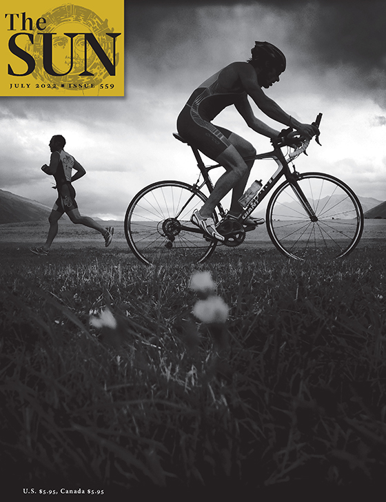 July 2022 cover of The Sun. A fit man is jogging off to the left, while another in bike shorts, tank top, and helmet is riding a bike off to the right. The photo was taken on the South Island of New Zealand on an overcast day.