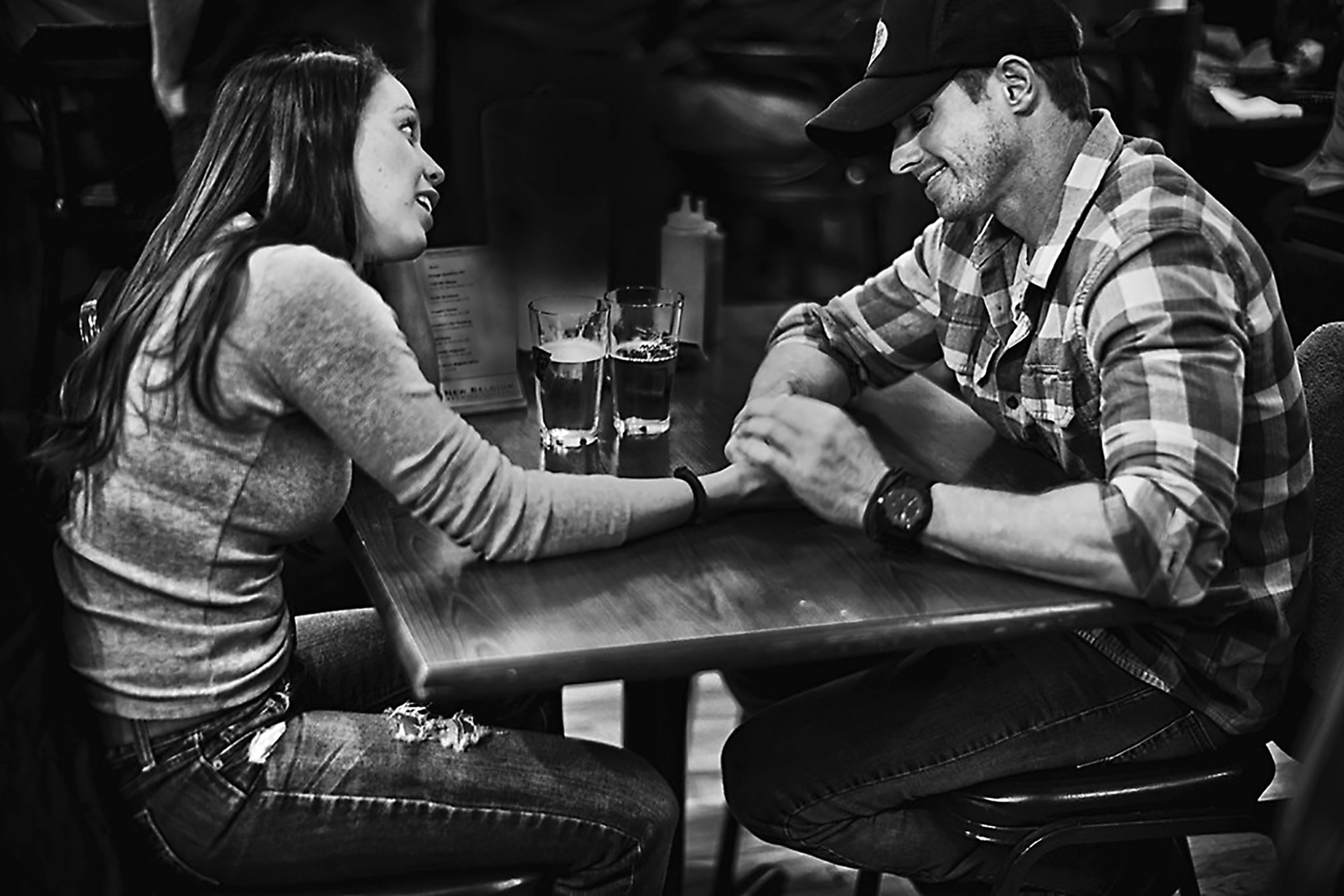 A woman and a man sitting in a restaurant holding hands across a table with two beers on it. They are both smiling, and she is looking at him while he is looking down. They are dressed casually in jeans and he has a cap on.