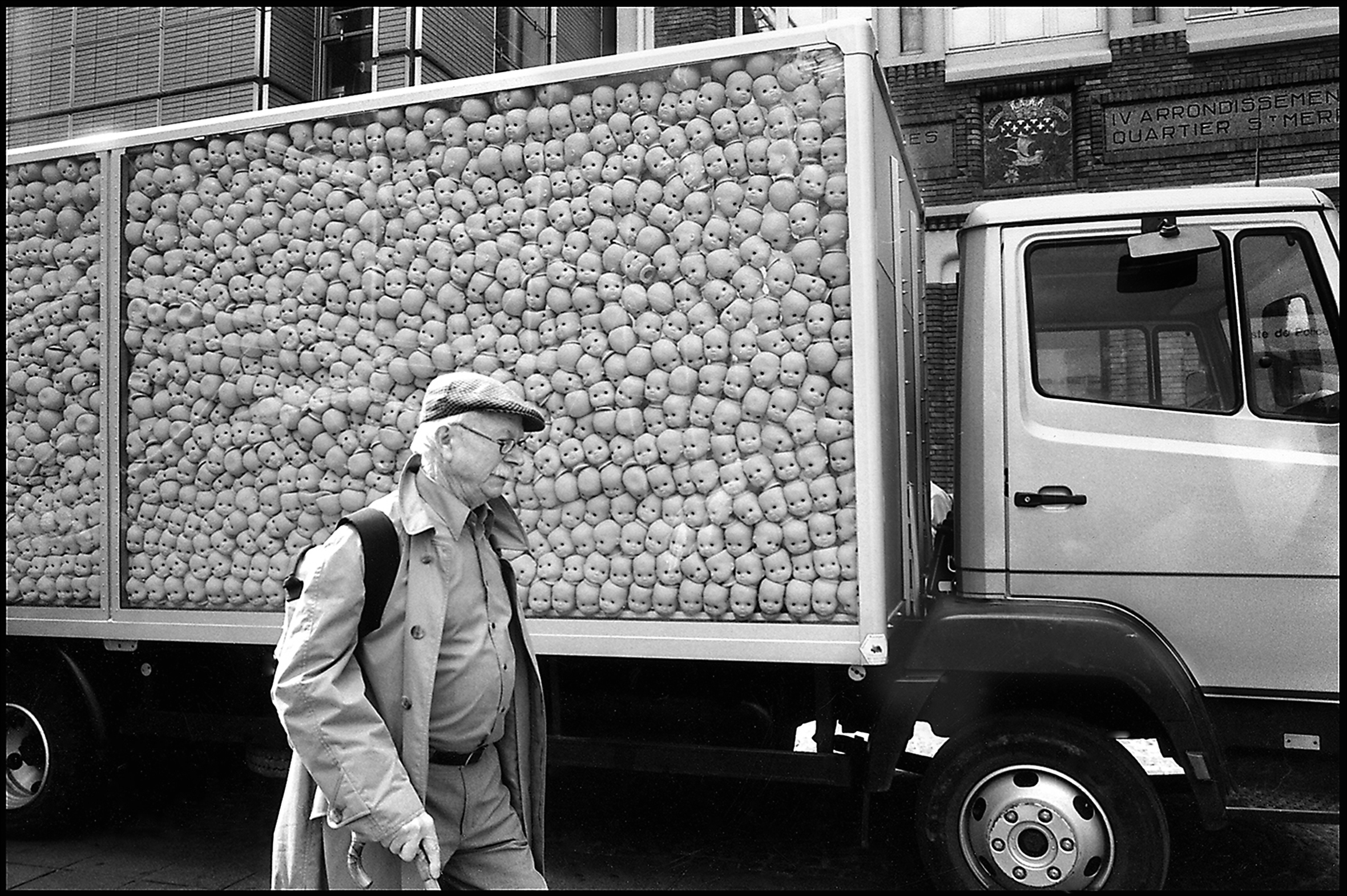 An old man with a cap and gray hair peeking out and a gray moustache is walking by the transparent side of a delivery truck filled with hundreds of identical small, white, bald, doll heads staring out.