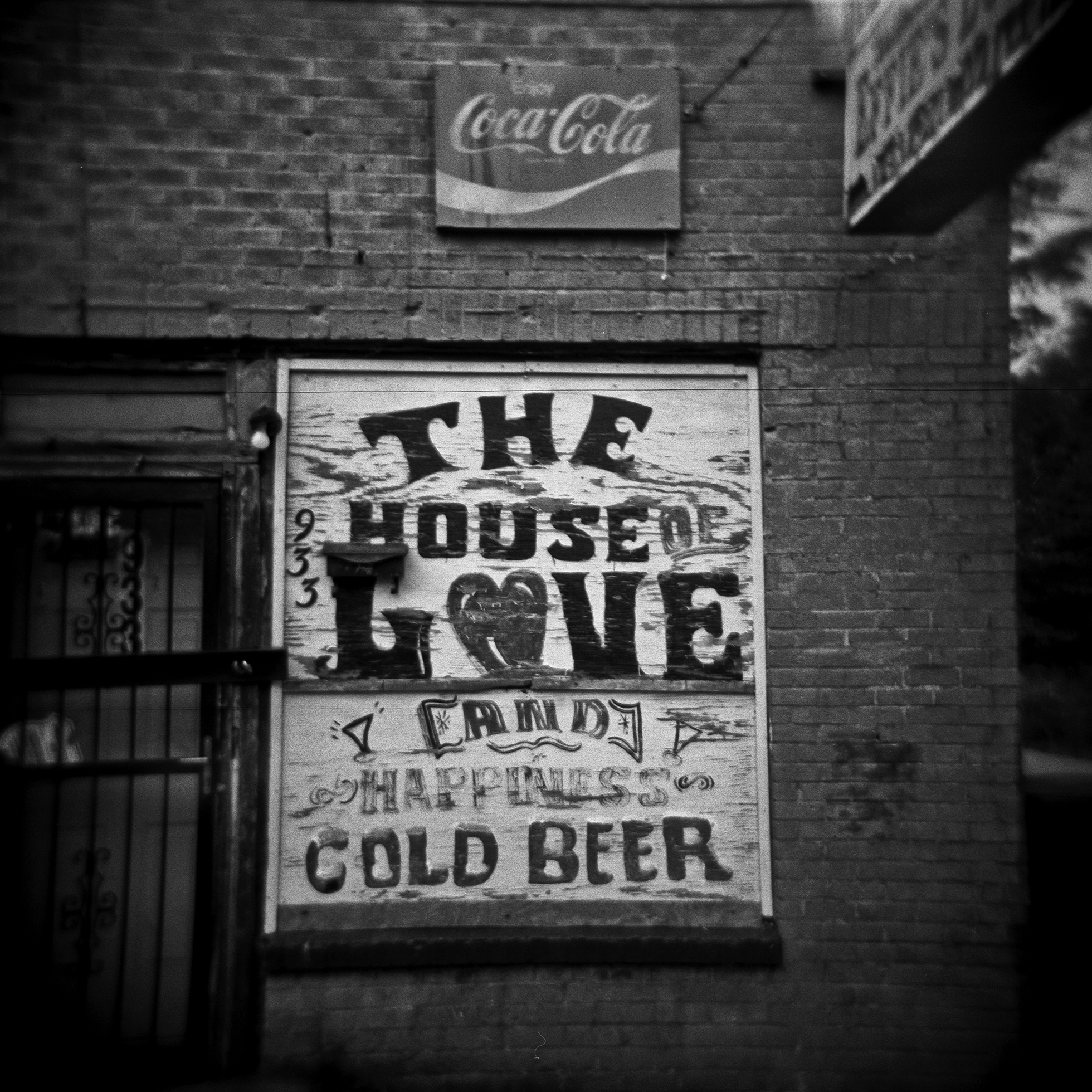 A brick building with an Enjoy Coca-Cola sign above a large sign next to the door that reads 933 The House of Love and Happiness Cold Beer.