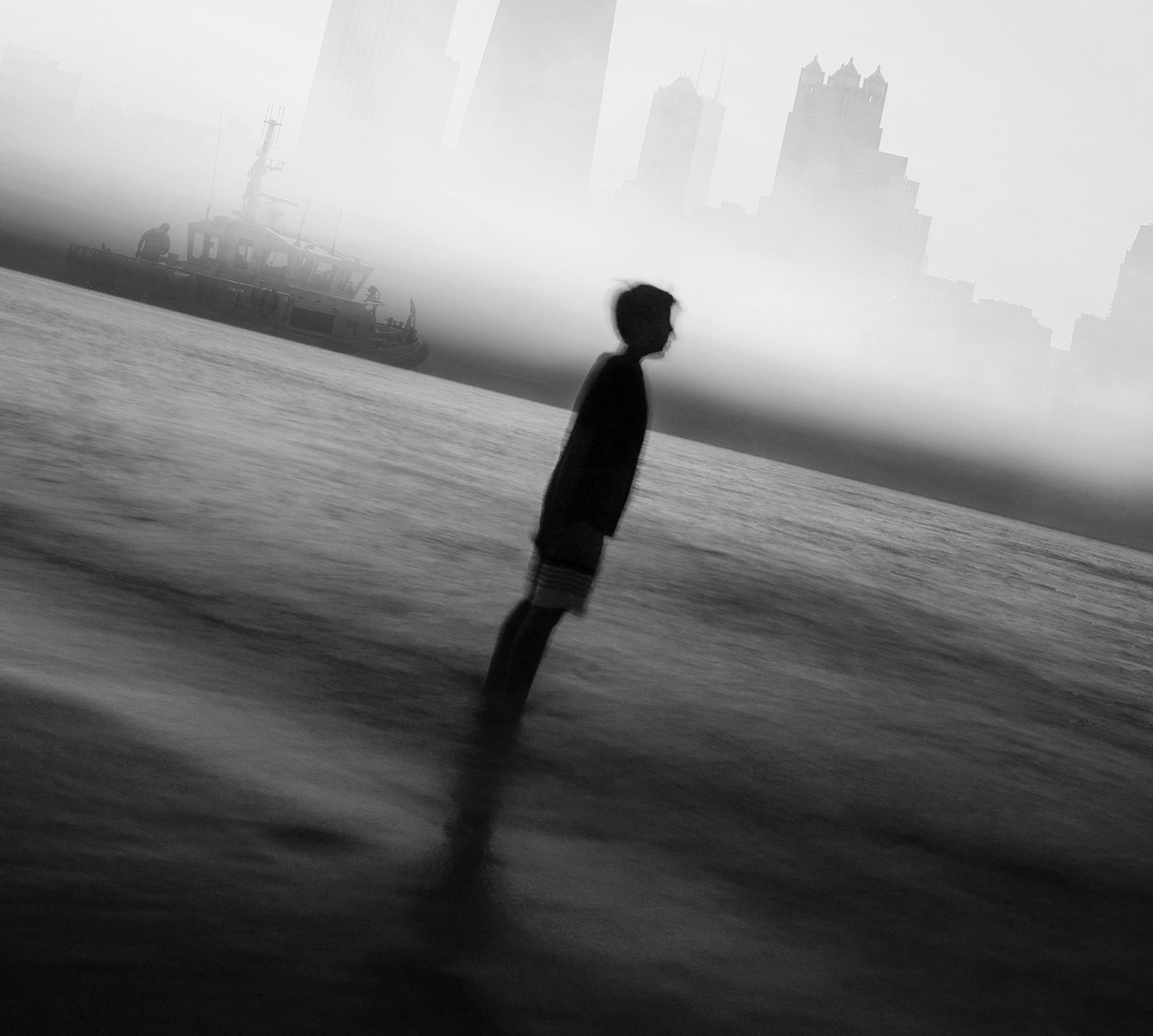 A boy in a T-shirt and shorts is standing ankle deep in a body of water. In the background is a boat, and beyond it are skyscrapers. The boat and buildings are shrouded in a mist, and the image has been rotated slightly to the right.