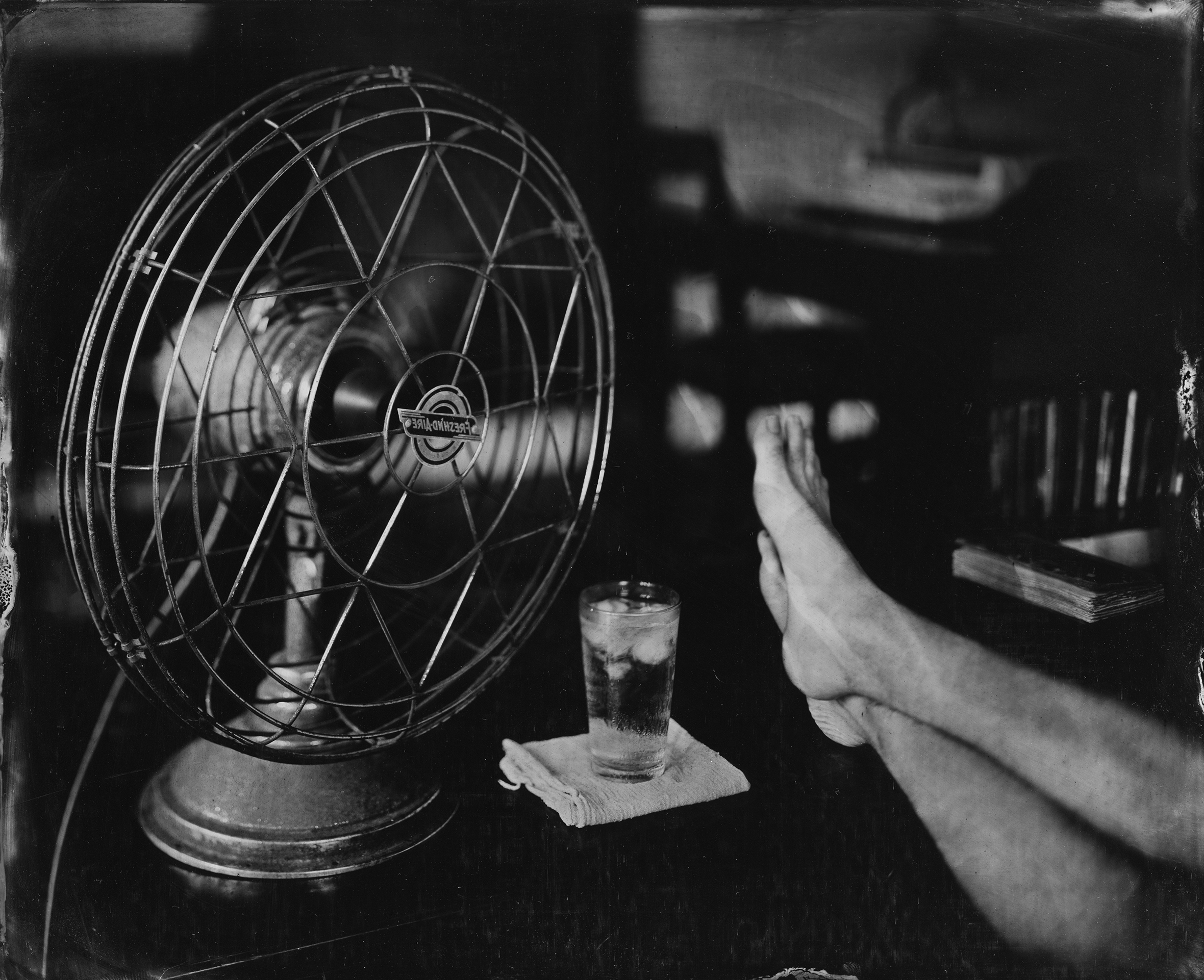 A desk with an iced beverage and an old-fashioned fan pointed at a seated man of whom is shown his bare shins and crossed feet.