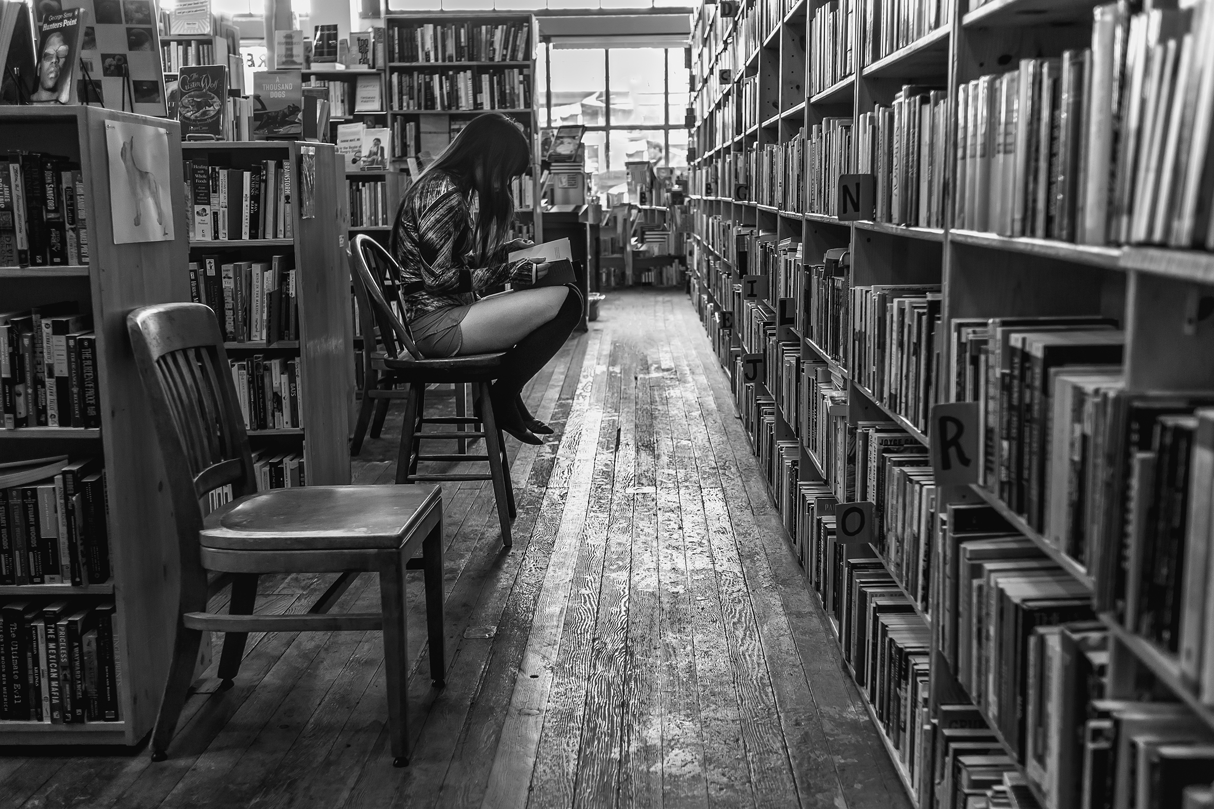 Looking down a main aisle of a bookstore with wood flooring to the large windows at the front. On the left, a girl reading a book is sitting on a wooden chair at the end of a bookcase facing a multitiered bookcase that runs the length of the long aisle.
