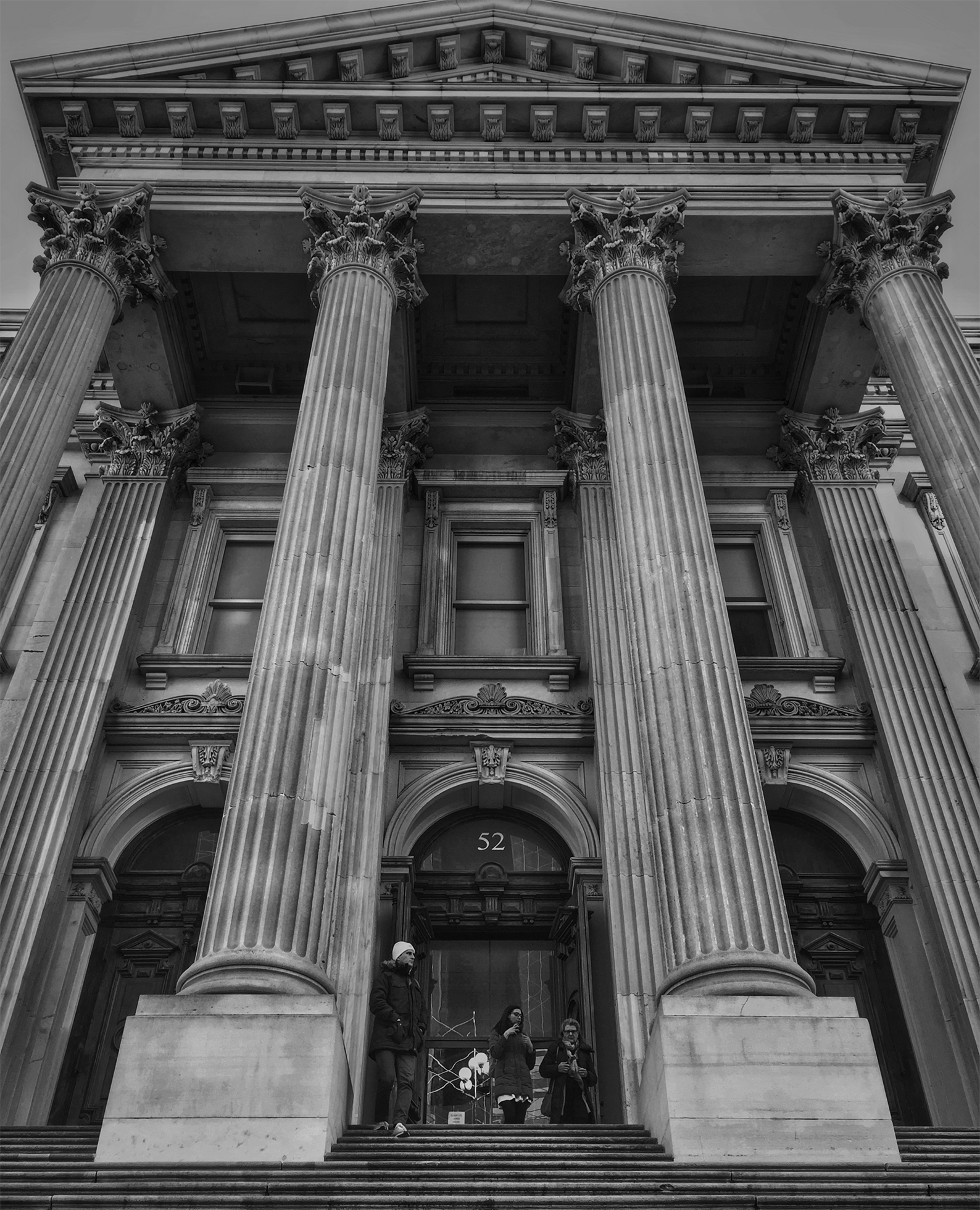 A looming court building designed with classic Greek architecture. A few people linger outside of the doors, looking small against the giant columns.