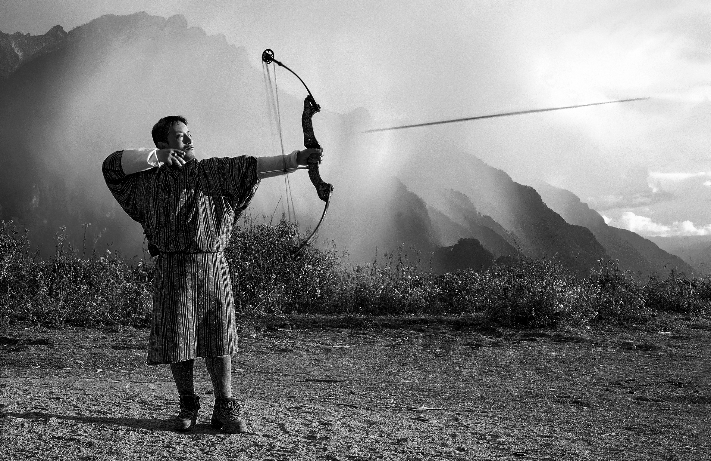 A Bhutanese archer stands among nature with mountains in the distance. He has just let loose an arrow.