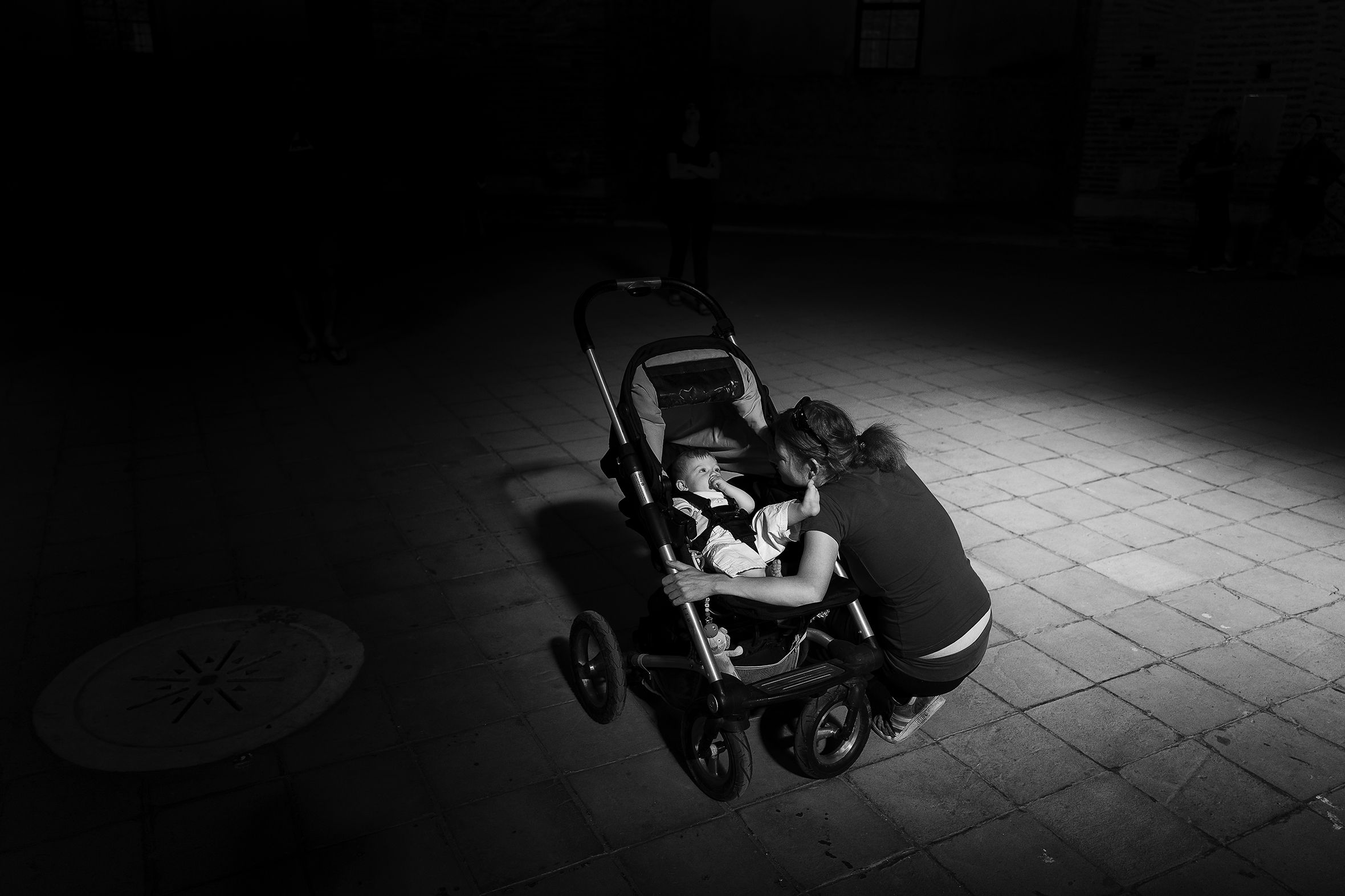 A woman bending down to look at a baby in a stroller. They are surrounded by a vignette, so it is only her and the baby cast in light.