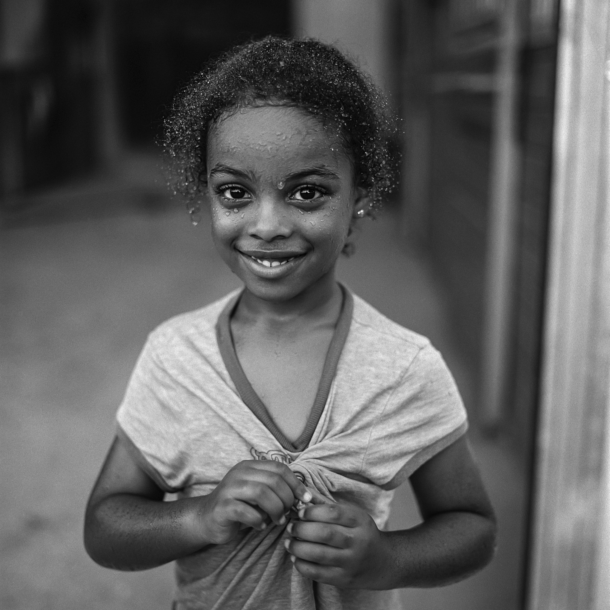 A young girl whose face and hair are wet smiles at the camera. She is standing in a barn and wearing a T-shirt that is dry that she crumples in her hands.