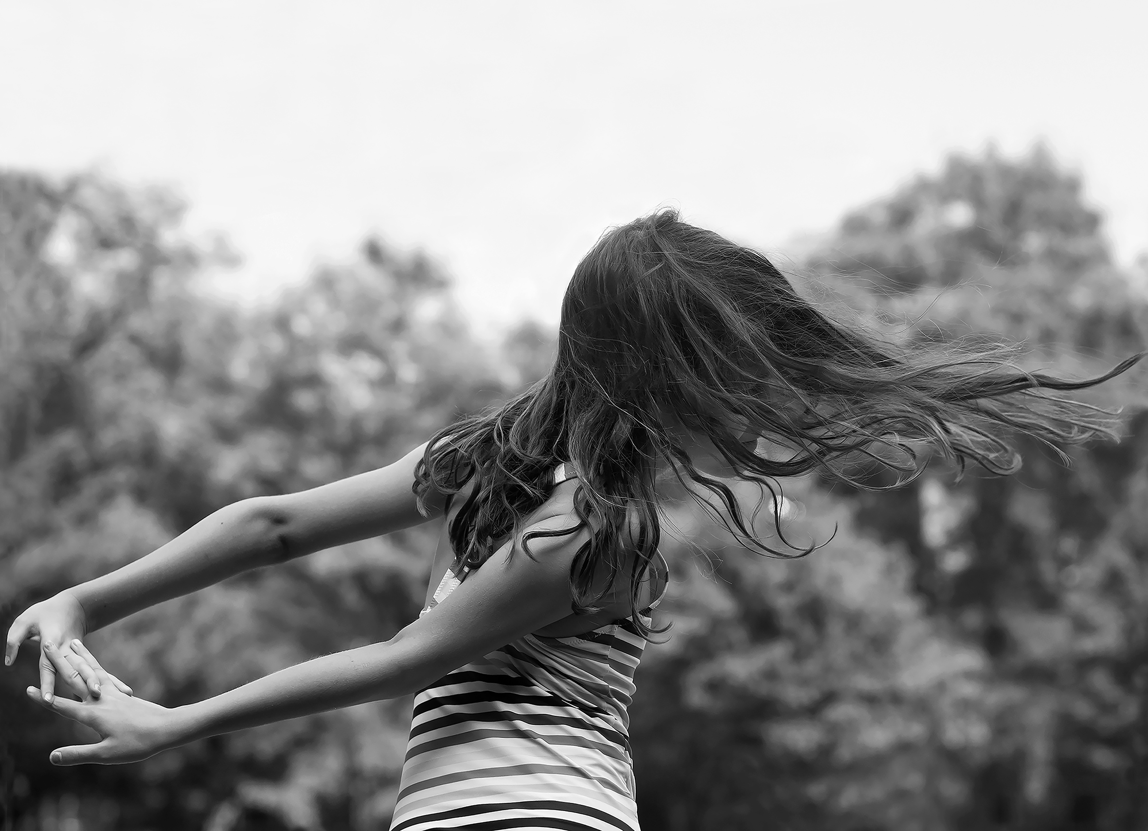 A young girl with long, brown hair is twirling around. Her arms are extended behind her and her fingers are interlaced, and her hair is flowing out as she twirls around outside with a backdrop of trees.