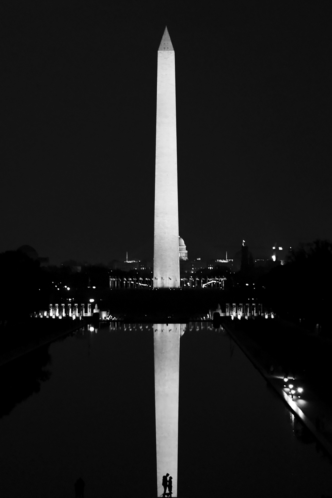 The Washington Monument at night. The image is black except for the monument itself, its long reflection to the bottom of the image, and lights of the city in the middle. At the bottom in the reflection, two silhouettes stand together.