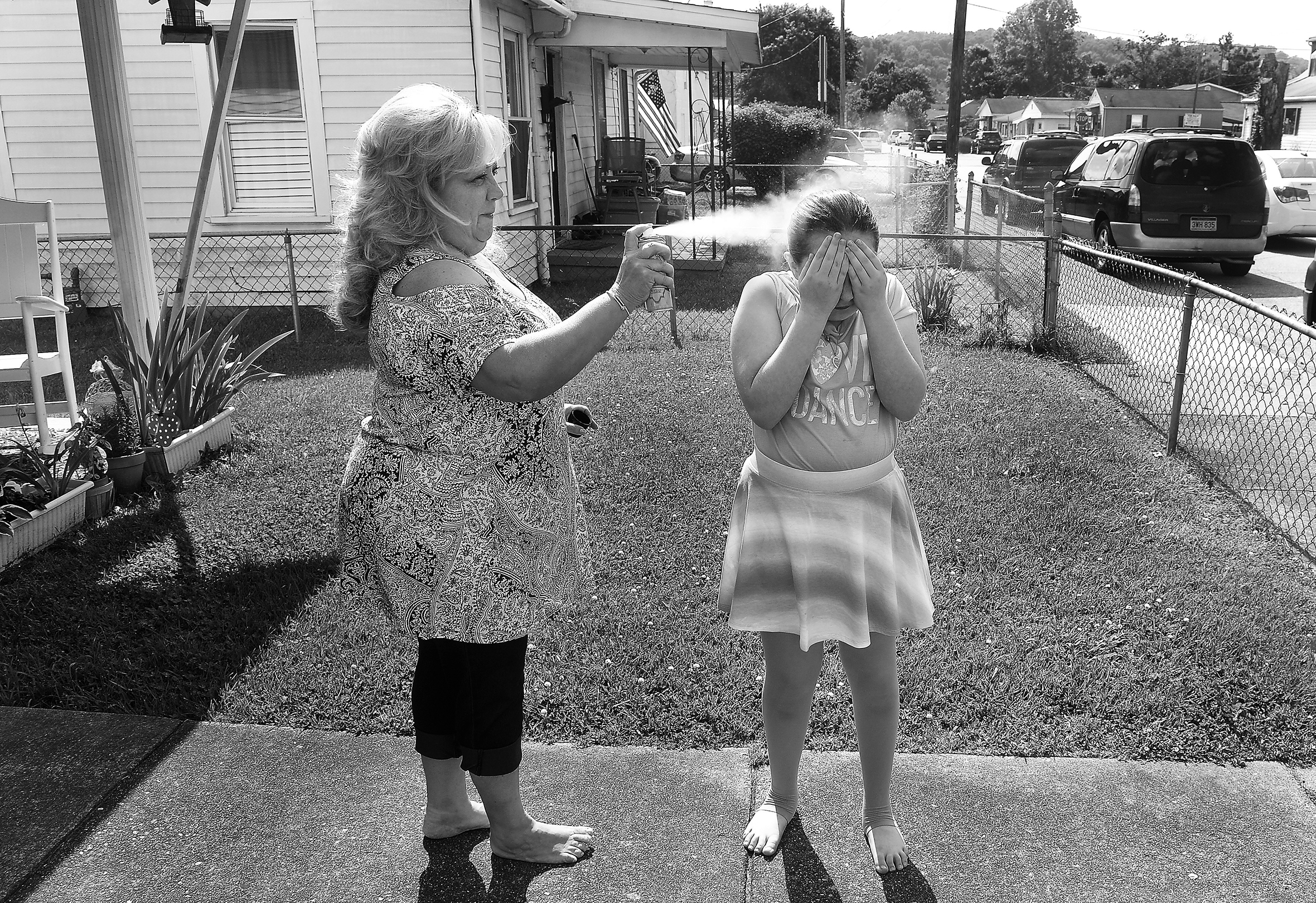 A woman with teased, blond long hair sprays something on a tween girl’s hair as the girl covers her face with her hands in a suburban front yard with a front porch and chain-link fencing.