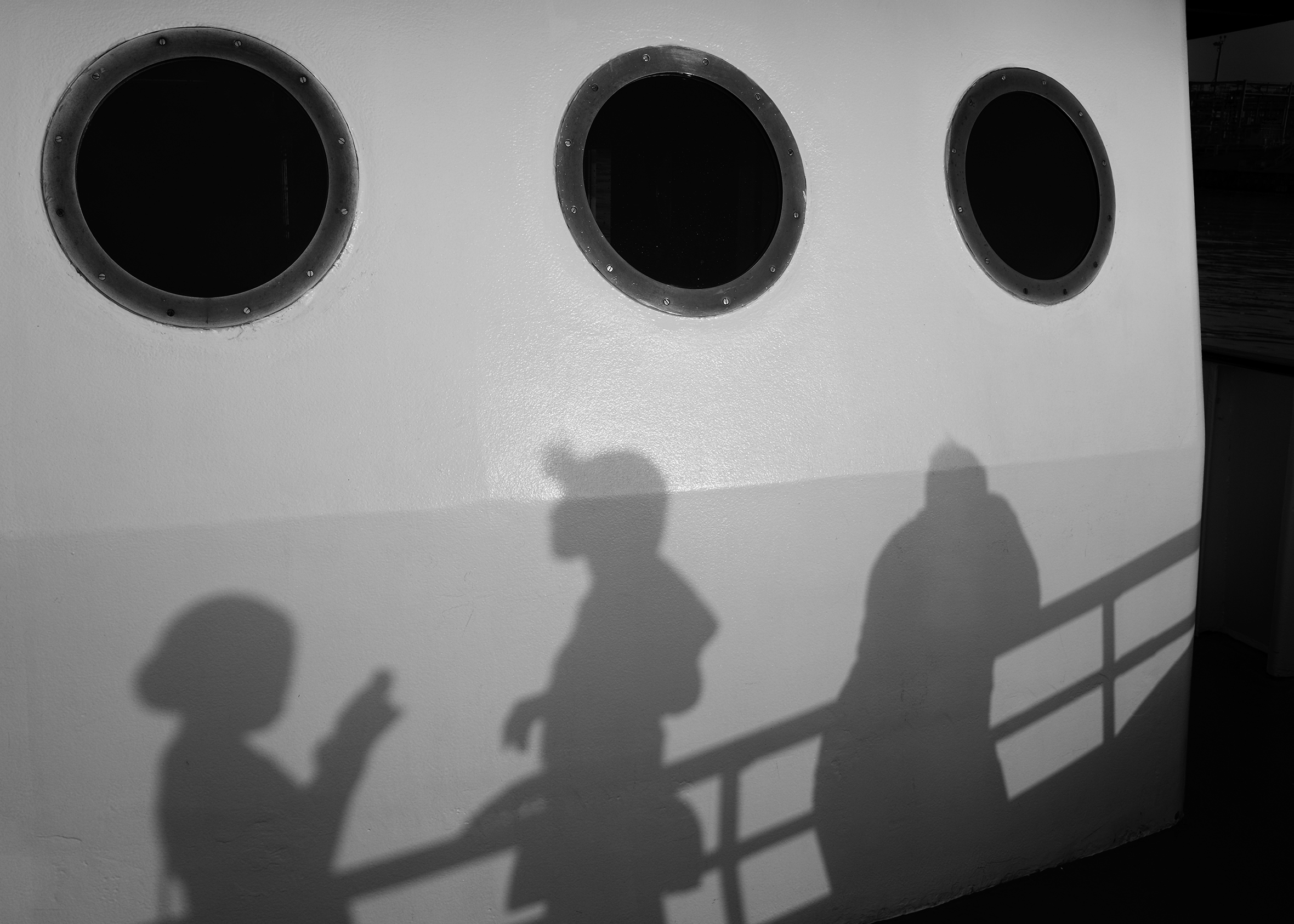 A close-up of three portholes on a ship. Reflections of three people against an open railing are on the side of the ship underneath the portholes.