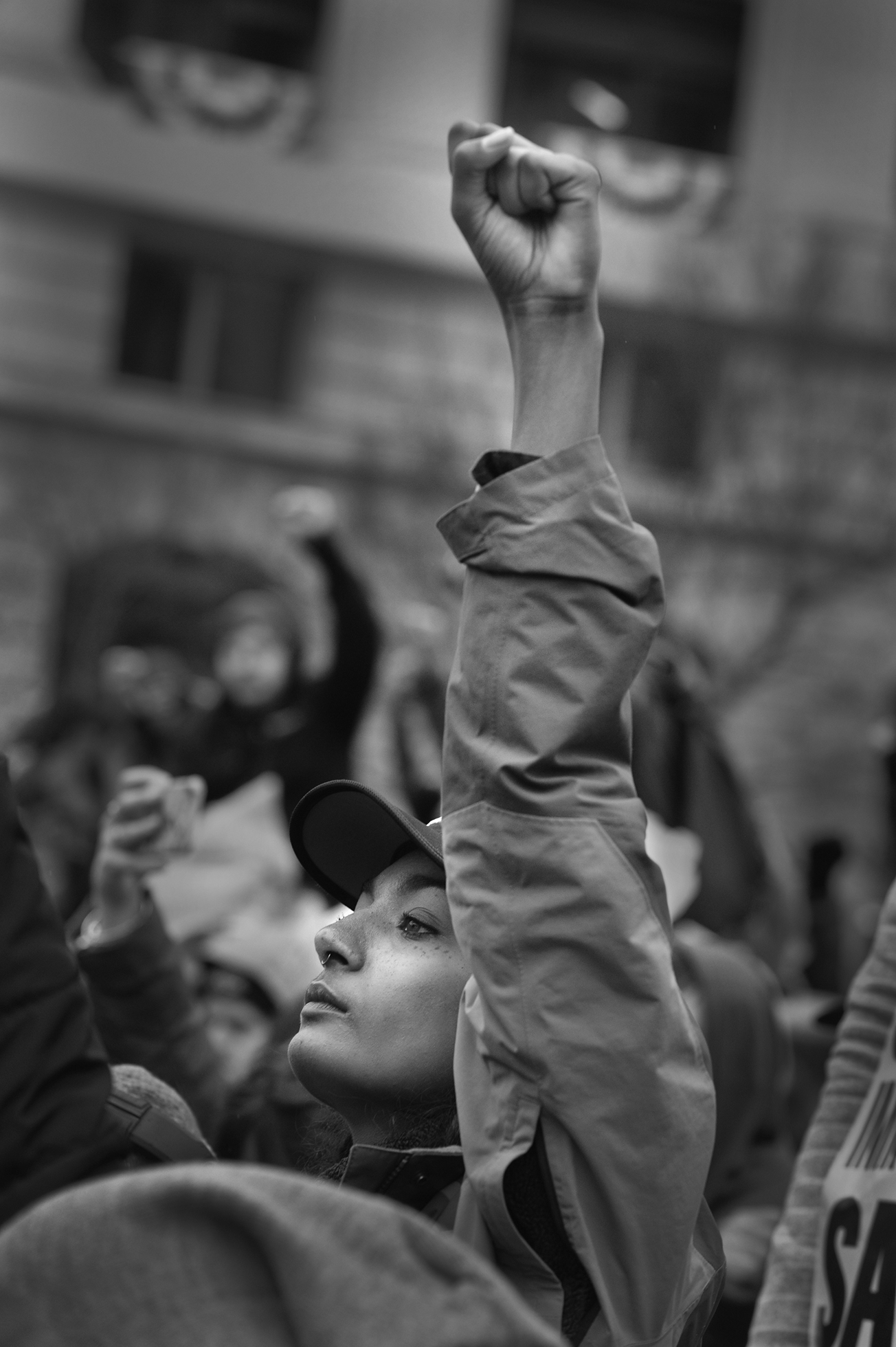 A determined looking woman facing left, protesting in a crowd on the street, with her left arm raised with her hand in a fist, wearing a baseball cap and jacket.