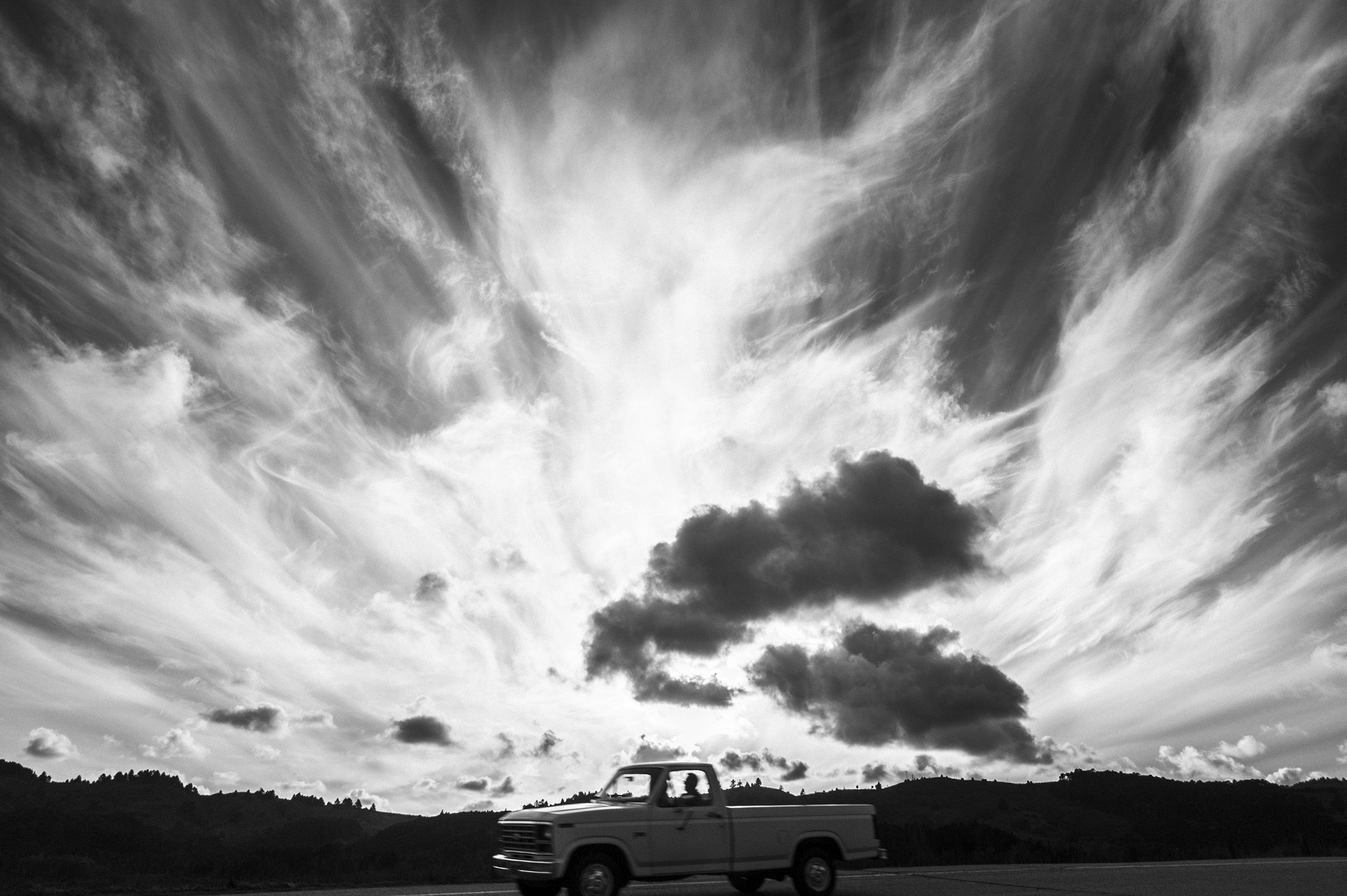 A wide shot of a man driving a pickup truck with low hills in the background. The largest and most striking part of the image is the sky. The clouds look flamelike.