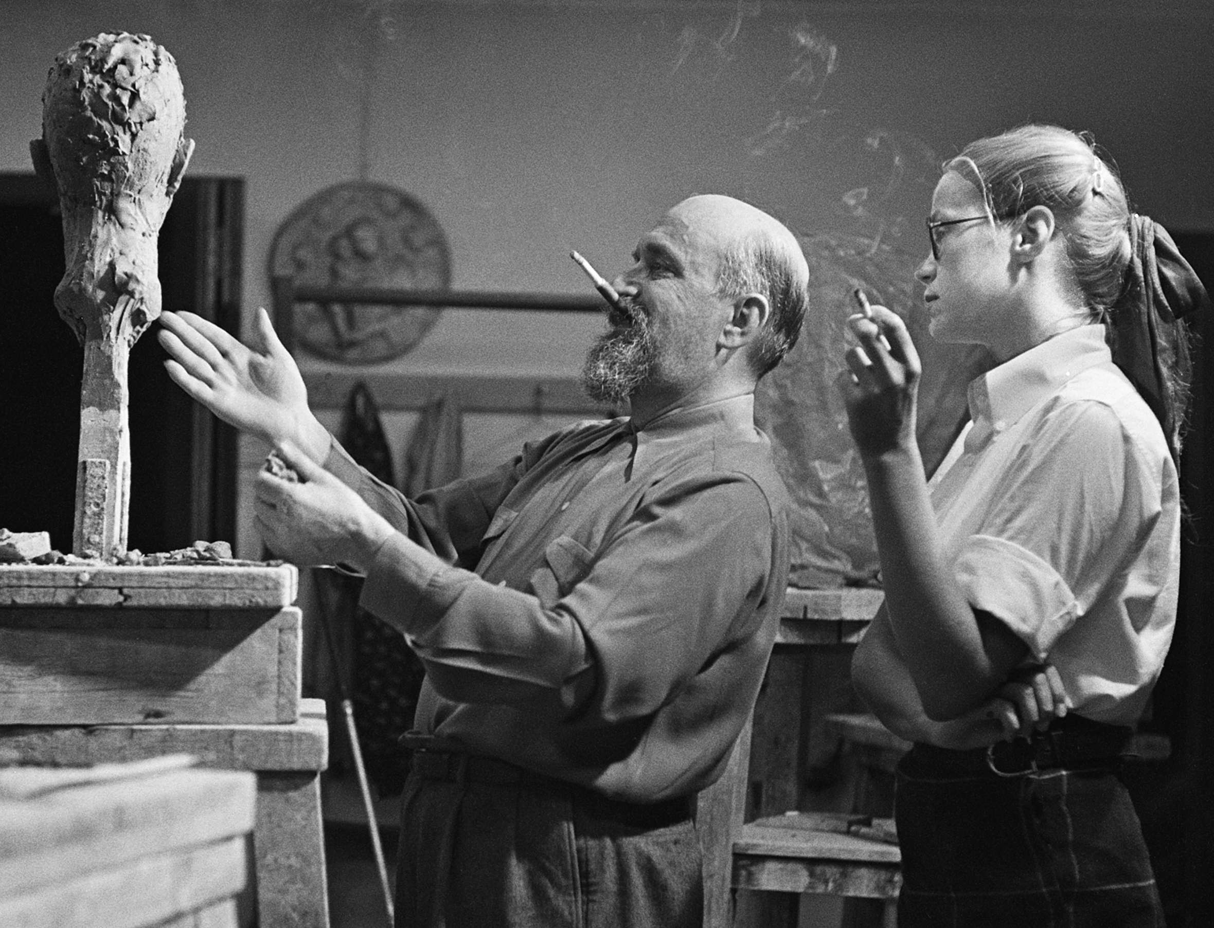 An older man and younger woman stand in an art studio or classroom discussing a clay bust on a pedestal. Both people are smoking. The man has a cigarette in his mouth as he gestures to the bust with both hands.