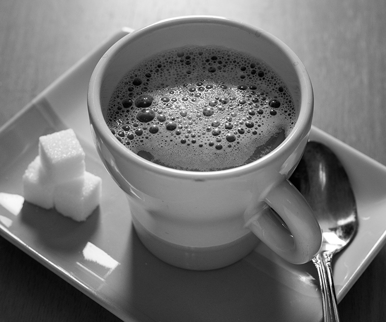 Close-up of a cup of coffee on a rectangular serving plate. Three sugar cubes are stacked to the left of the cup and a teaspoon is placed to the right.