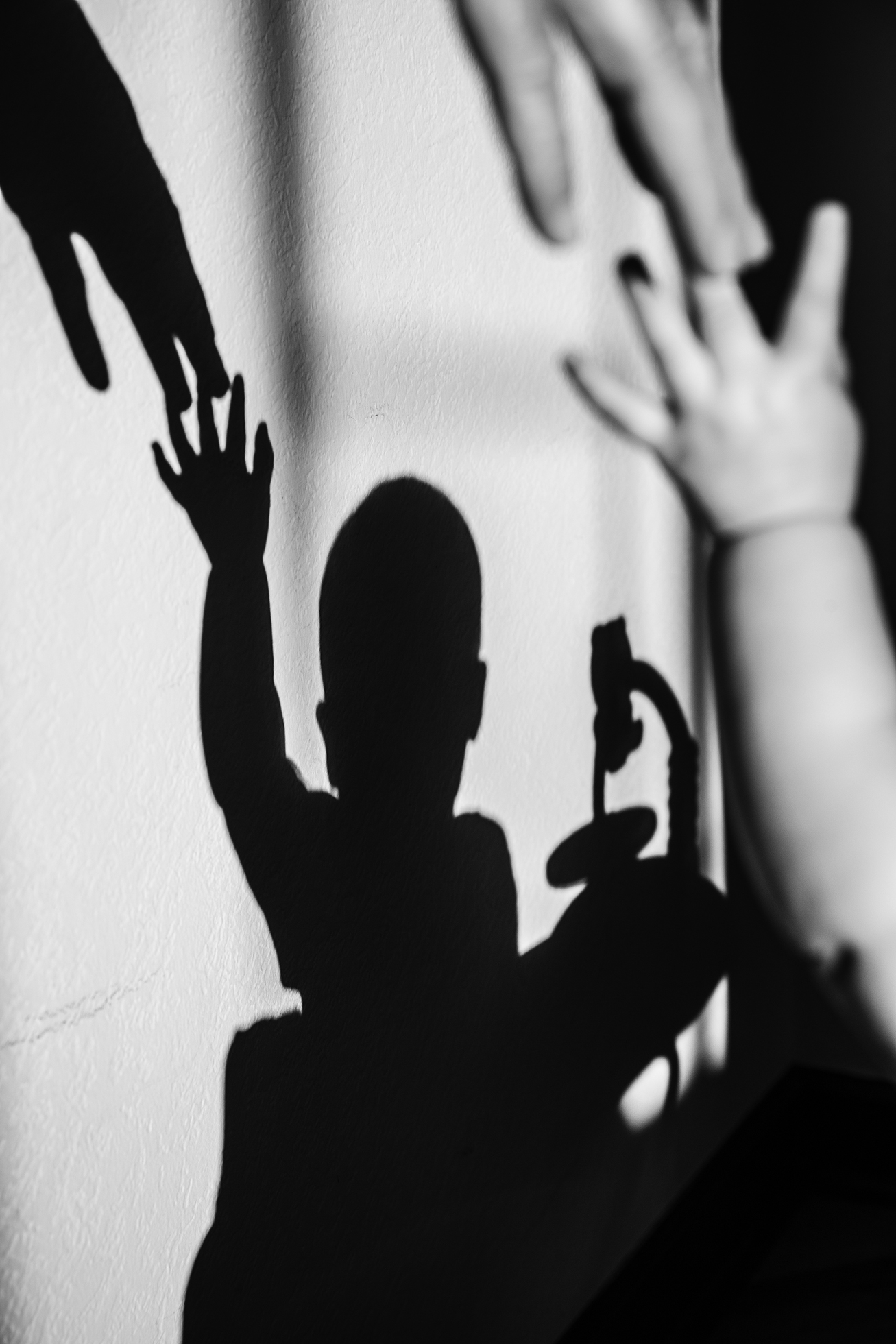 A shadow on a wall of an adult’s fingers reaching down and touching the fingers of a child’s upstretched arm. The shadow shows the child’s head and torso as well.  On the upper right is the actual adult’s fingers and child’s arm and hand.