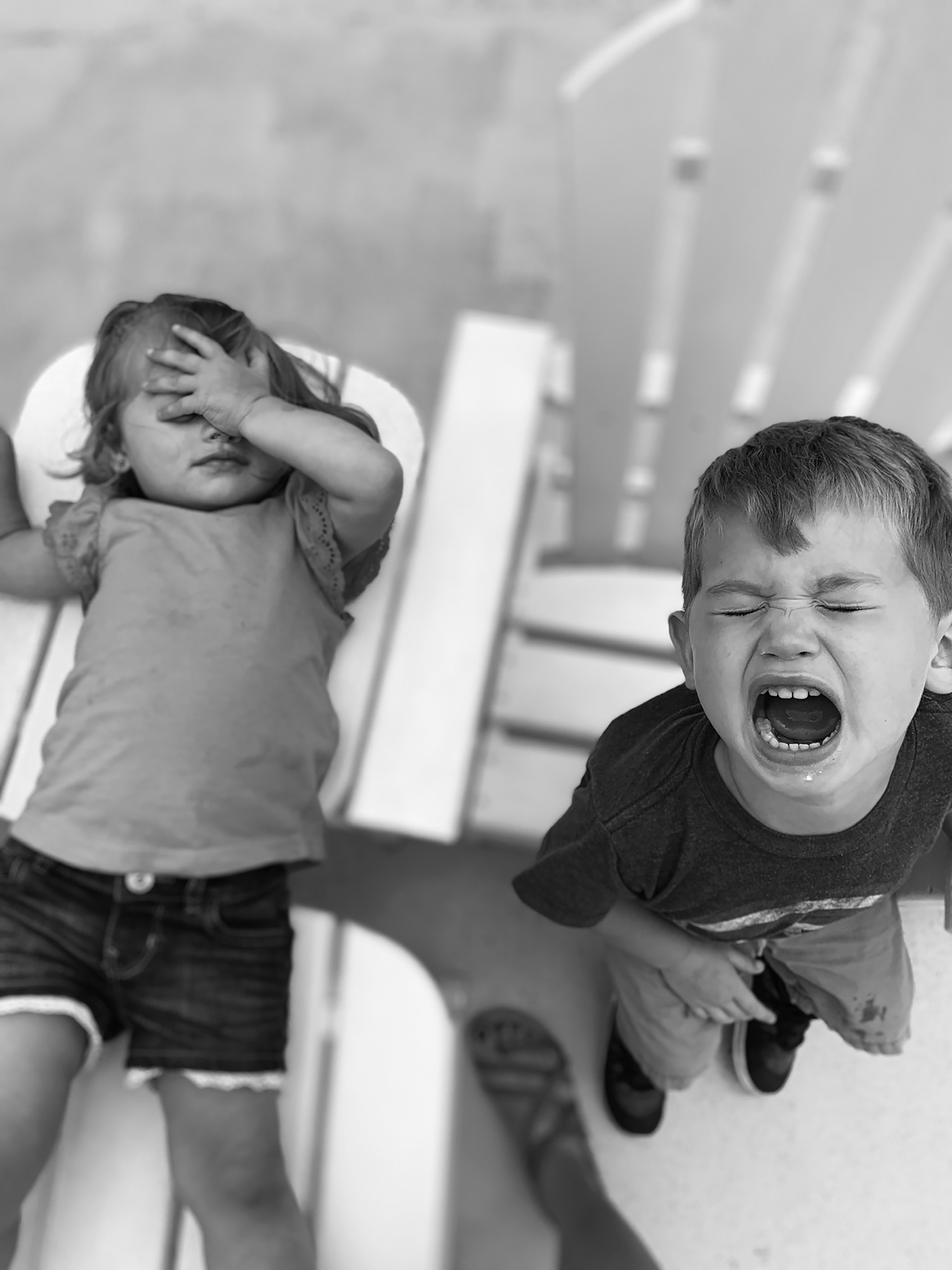 A young girl lays back on an outdoor chair with one hand over her exasperated face as a young boy stands next to her crying loudly from the looks of it, and in between them, one adult woman’s sandaled foot adds context.