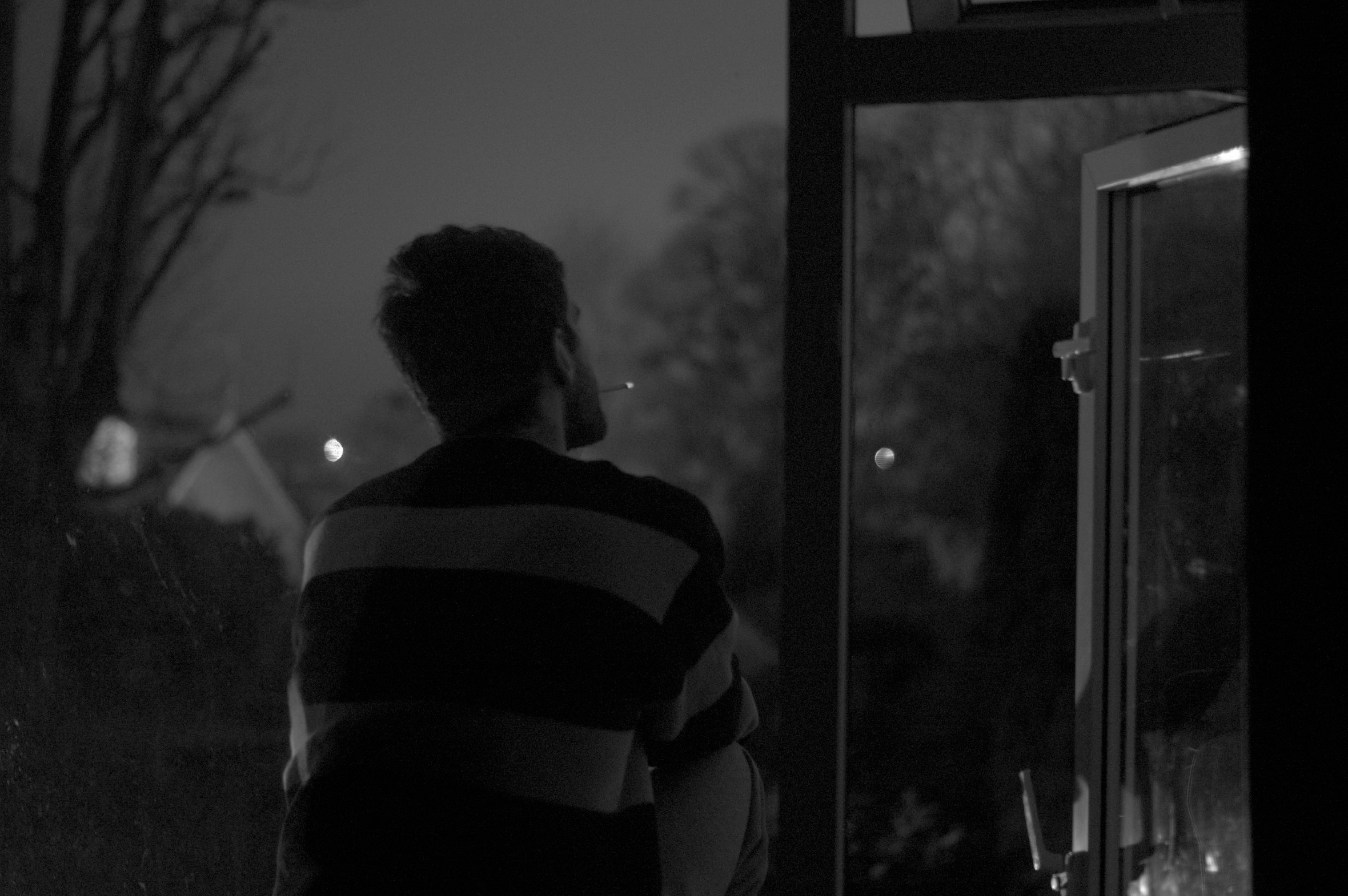 A young man sits and smokes outside of a home in a suburban neighborhood at night.