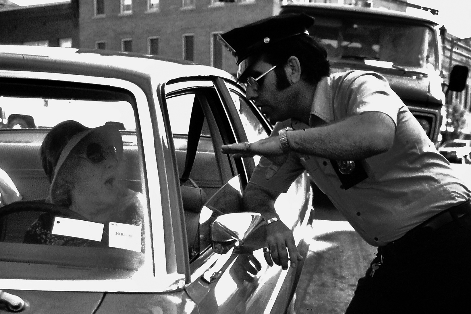 A police officer with sideburns and dressed from the 1970s speaks with an older woman in her car through the driver’s side window as he leans against the car door.