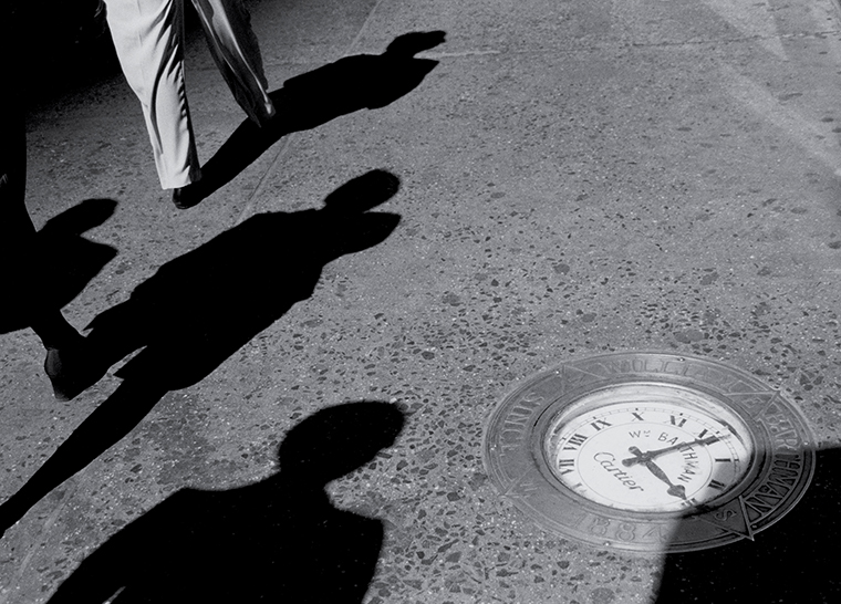 A clock set in a sidewalk at 3:01 PM with passersby evidenced by their shadows.