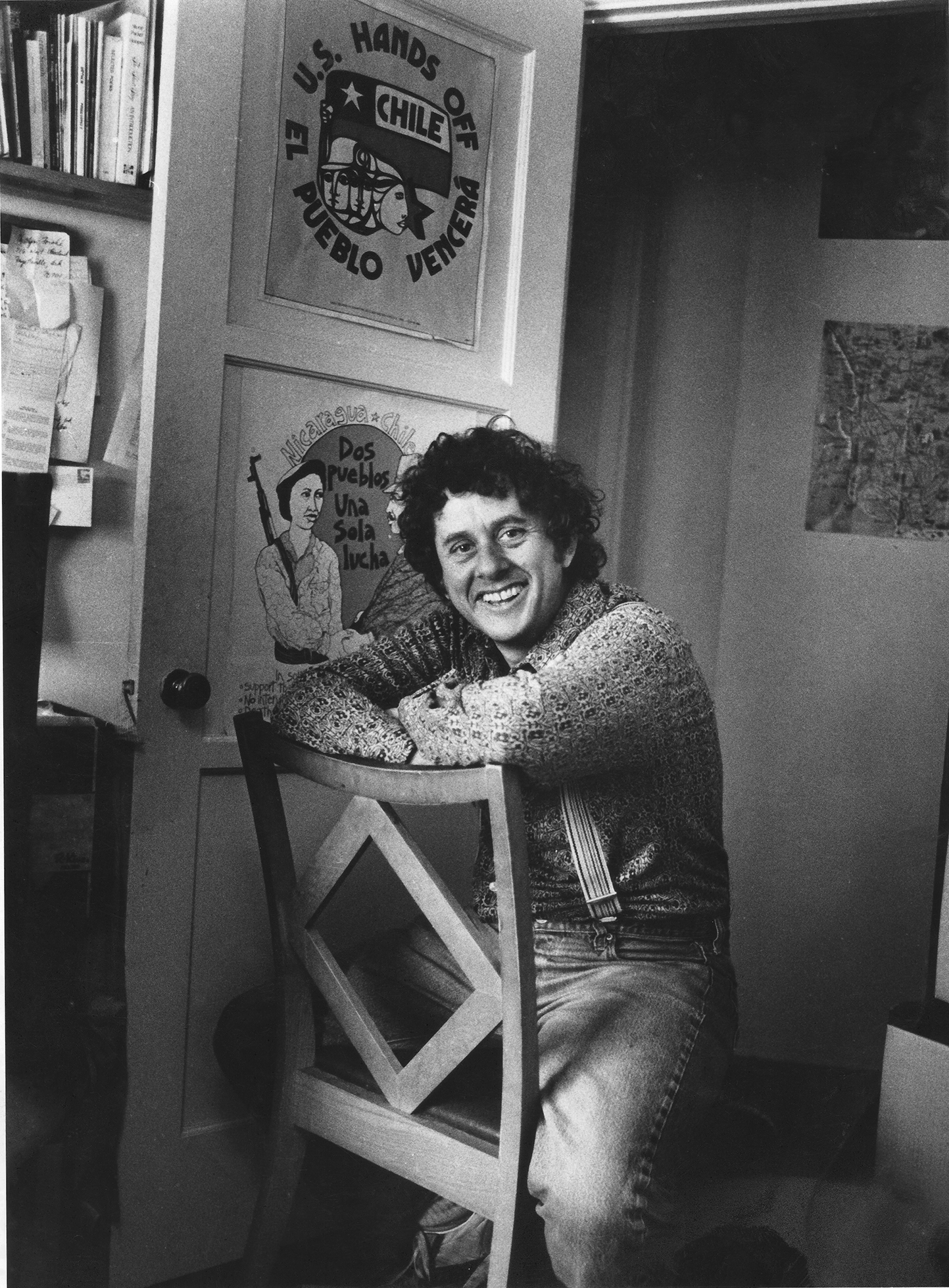 Steve Kowit is relaxed and smiling broadly sitting backward in a chair with his arms resting on the top. He is wearing jeans and a sweater and suspenders.
