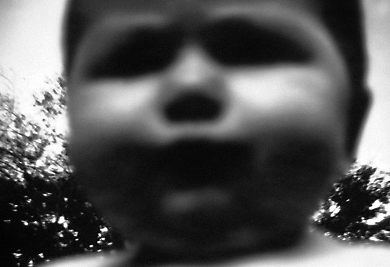 Close-up of the nude upper half of a toddler standing in front of bushes. The toddler’s image is blurry and the toddler appears inordinately large.