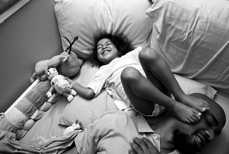 Bedtime shenanigans with Carlos Richardson and his daughter Selah. She is holding a stuffed animal and smiling while on her back on the bed with her feet on top of her father’s smiling, turned-to-the-side face.