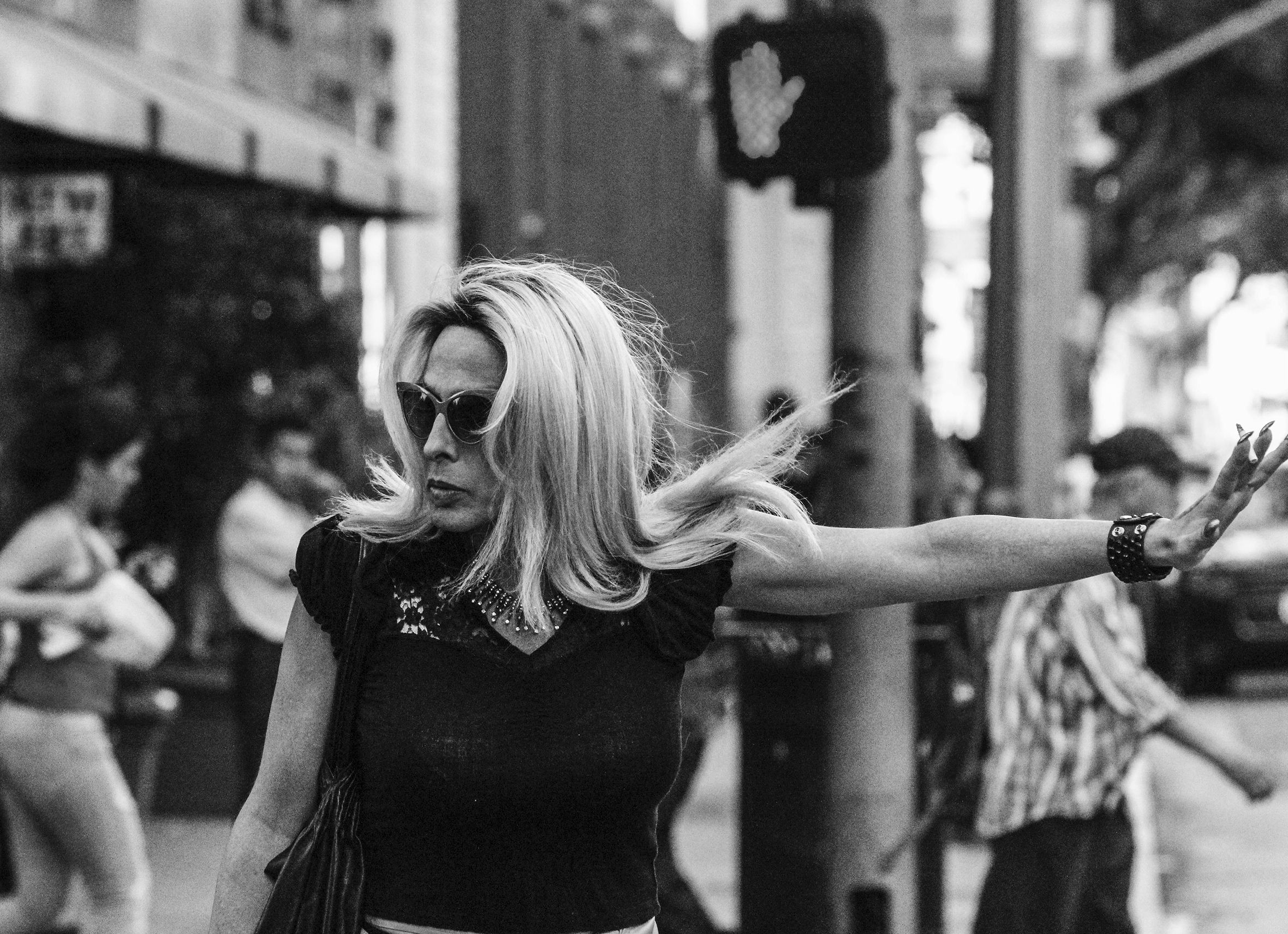 A woman in sunglasses with a resolute look stands facing the camera but looking right and extending her left arm straight out to the side with her hand raised signaling stop, while on a city street in front of a pedestrian signal of an upraised hand.