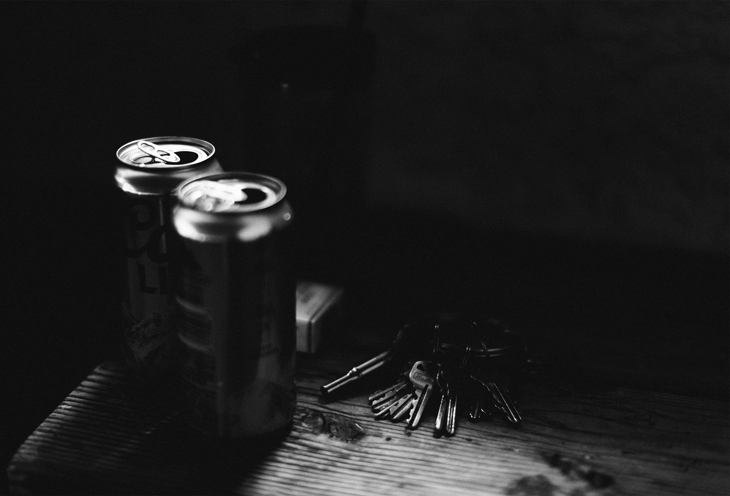 The corner of a wooden table upon which are two open beer cans with a pack of cigarettes peeking out from behind them and a plain metal keyring with many keys. The table is in a darkened room and the objects on the table are mostly in shadow.