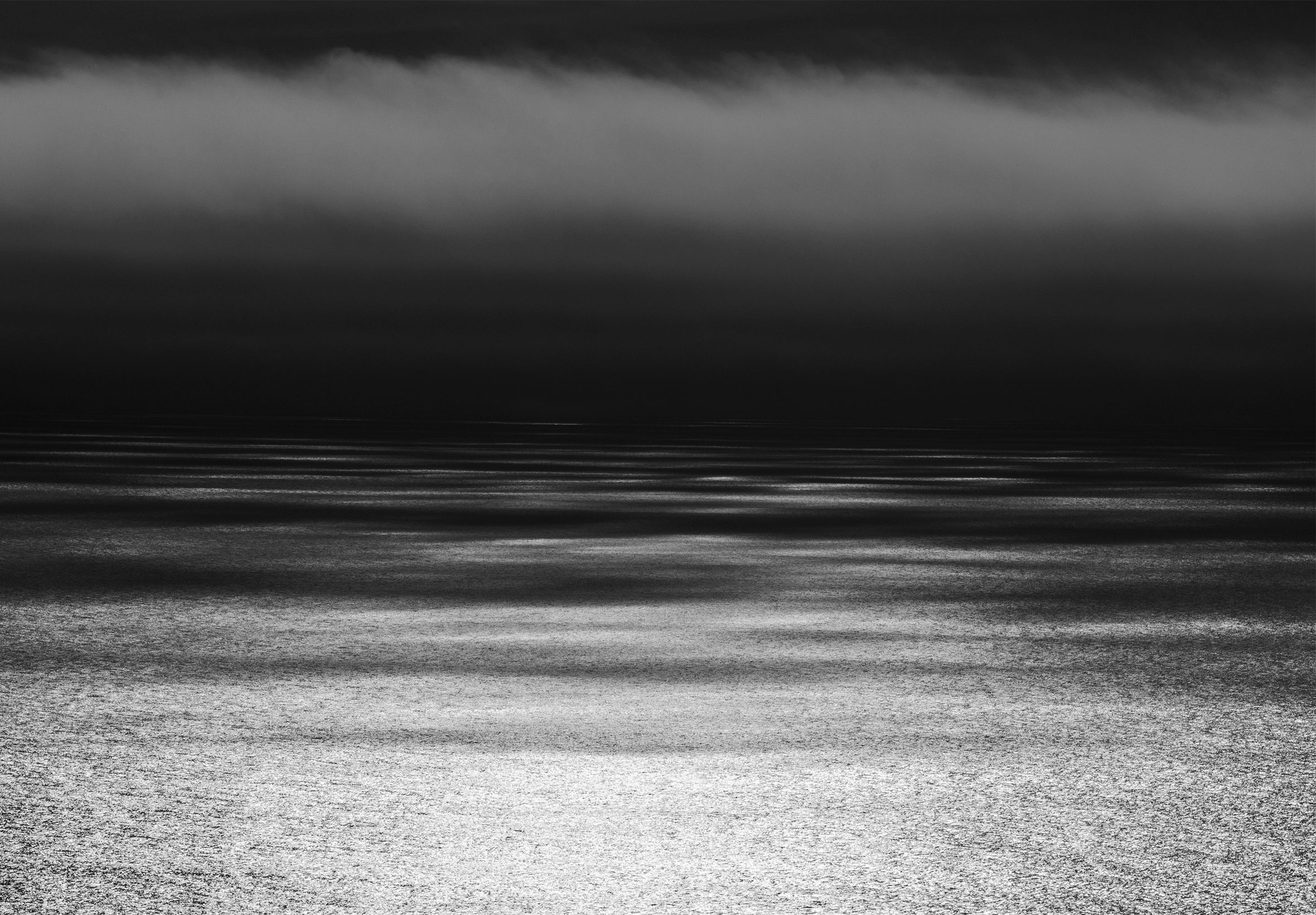 View from the beach of the water to the horizon with dark clouds peeking down from along the top edge of the image. The image is dark with light rippling on the water.