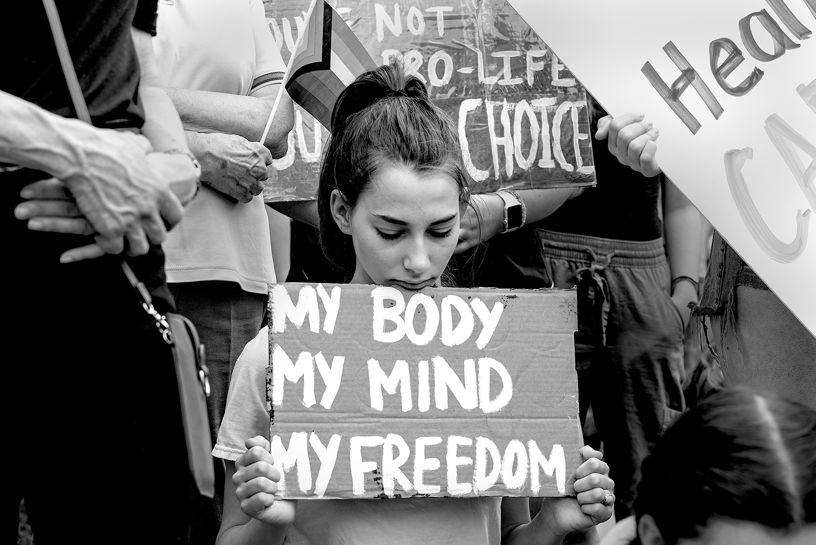 A young woman at an abortion rights rally looks down at the small cardboard sign she holds that says “My Body, My Mind, My Freedom” in white hand-lettering.