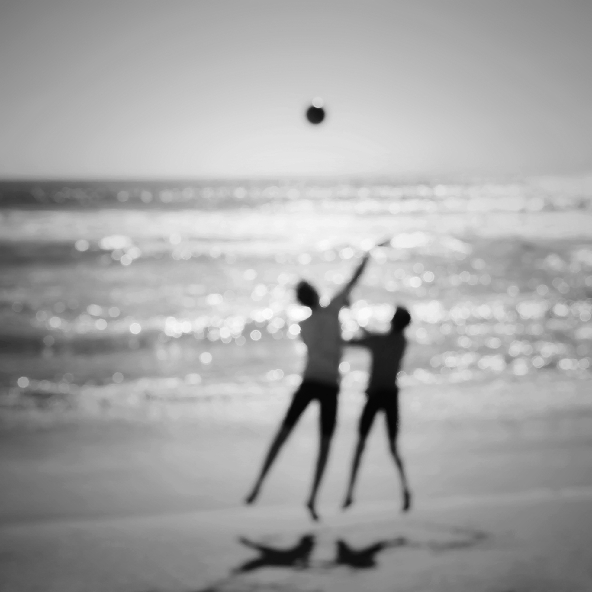 A blurry image of two boys in midair on a beach in Mexico. Both boys are positioned above the sand as the taller boy tosses a ball up in the air and the shorter boy outstretches his arms to catch it. The Pacific Ocean glistens to the horizon.