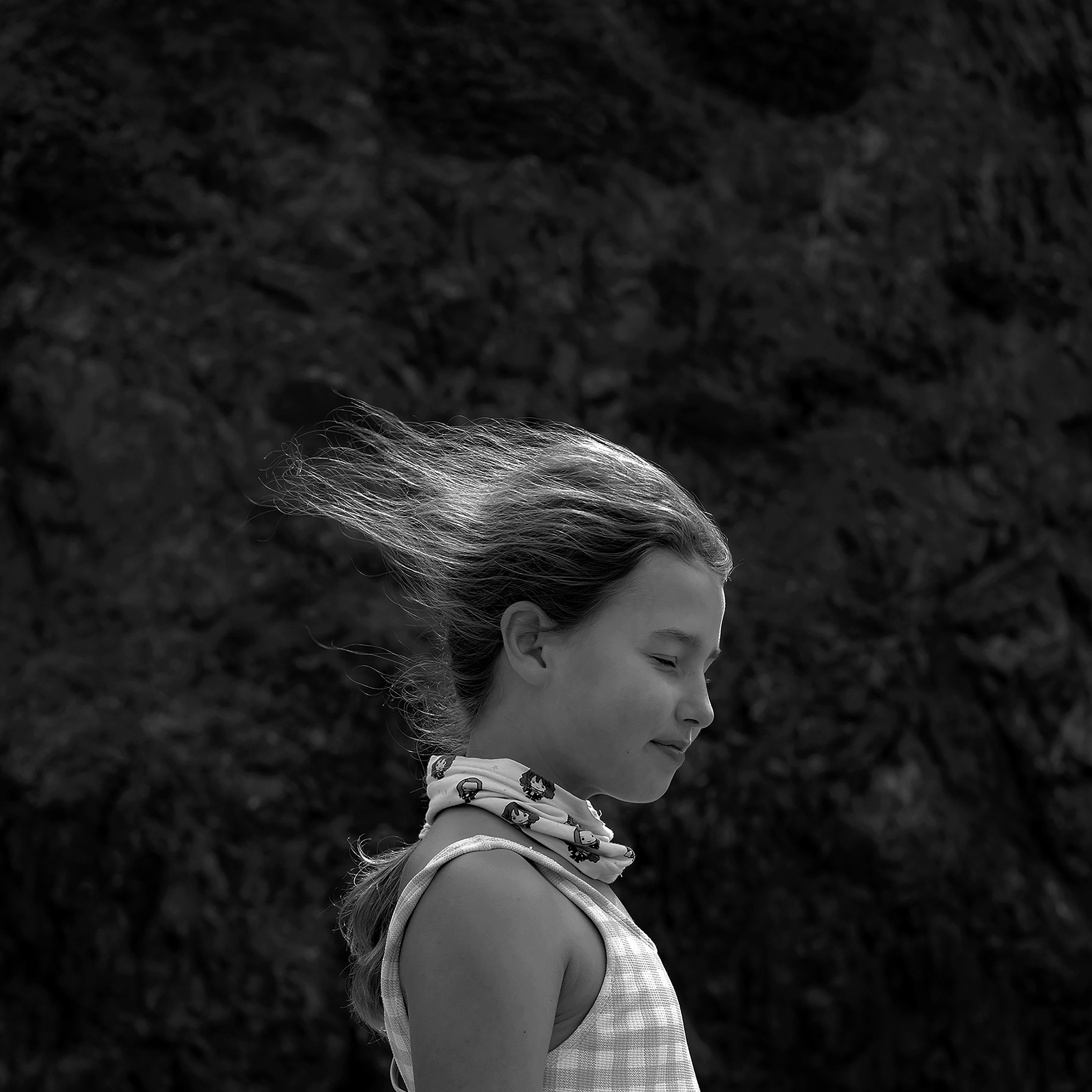 A young girl closes her eyes and mouth and smiles slightly as loose strands of her hair stream straight back in the wind. She stands in profile in front of a dark, rocky background on a windy day at Oregon’s Hug Point in 2020.