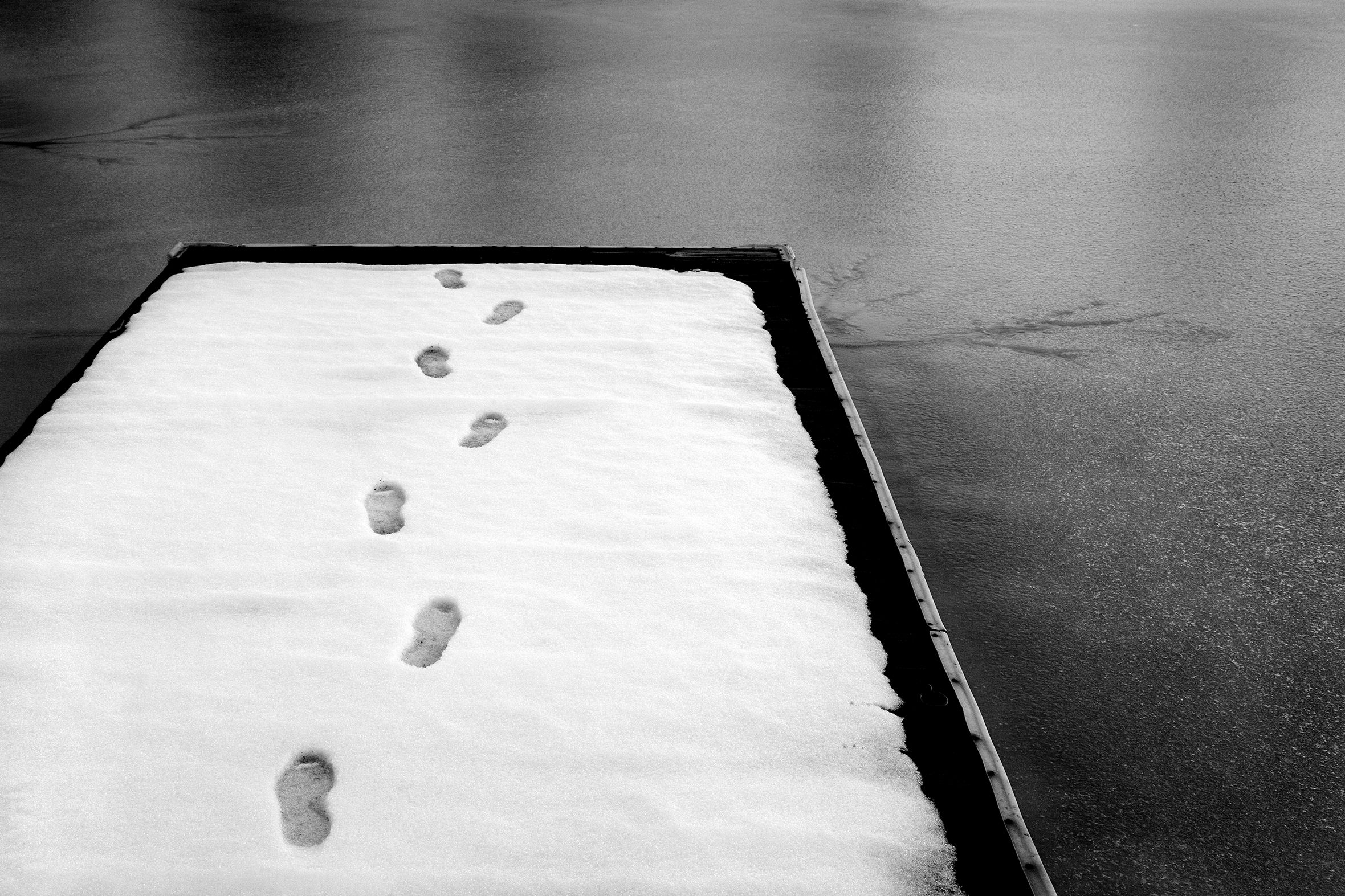 A small, snow-covered pier extends above frozen water. In the snow is one person’s footprints that walk the length of the pier and seemingly off the edge as the snow is otherwise undisturbed.