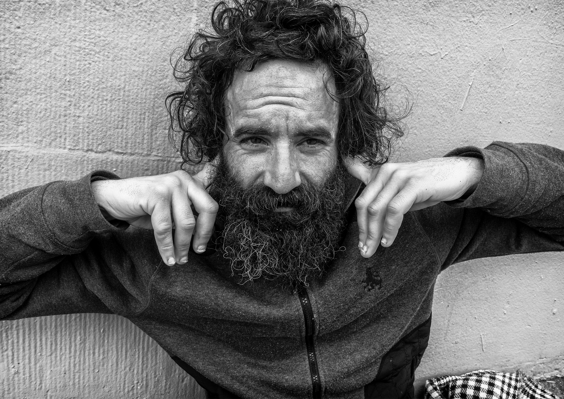 Delson sits on a sidewalk in front of a wall in San Francisco in December 2019. He has wild hair, a mustache, and a beard, and he looks ahead with a finger in each ear. He is wearing a light jacket.