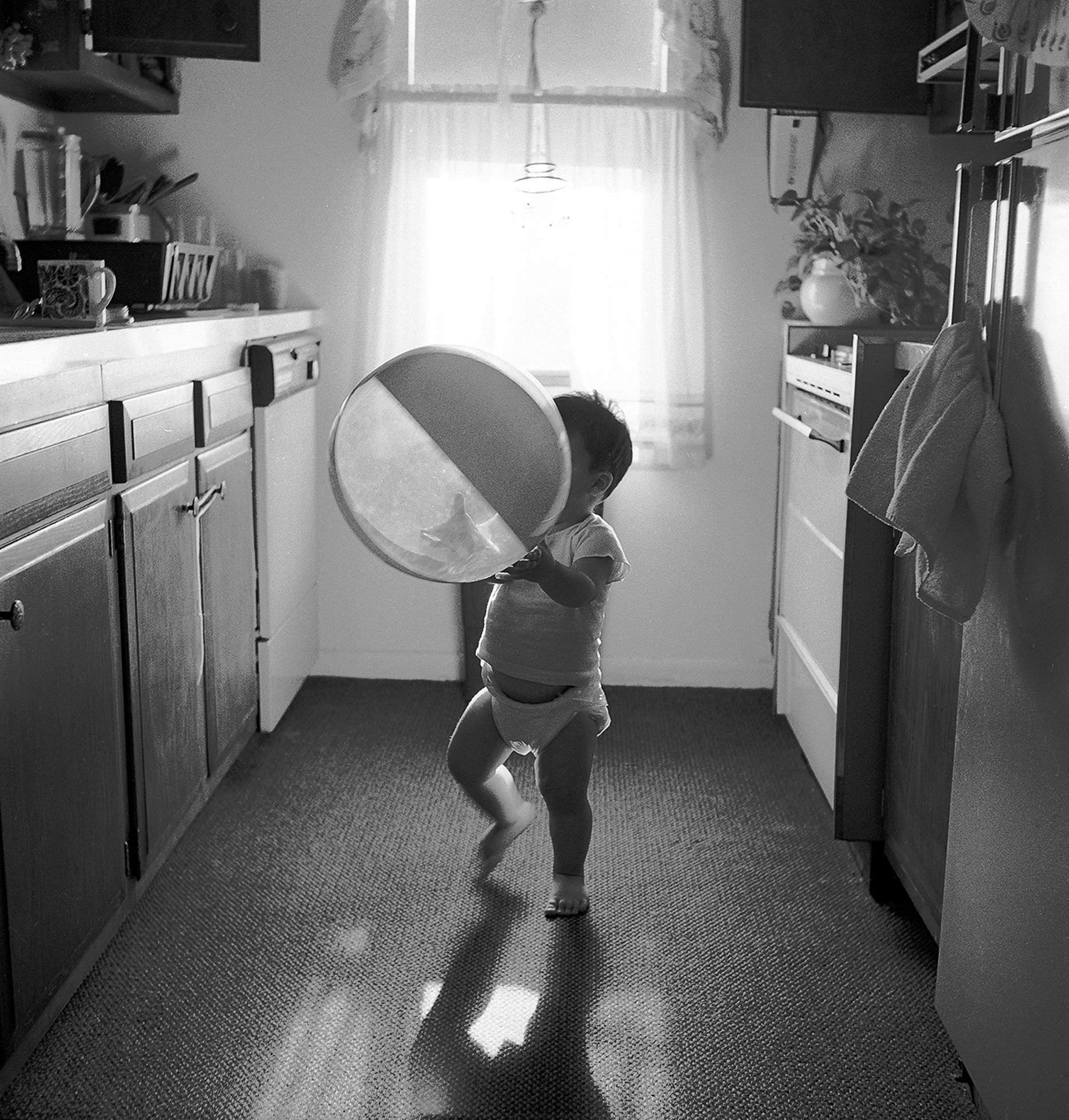 A barefooted toddler in a diaper and undershirt stands in a clean, bright, galley kitchen holding a large bowl.