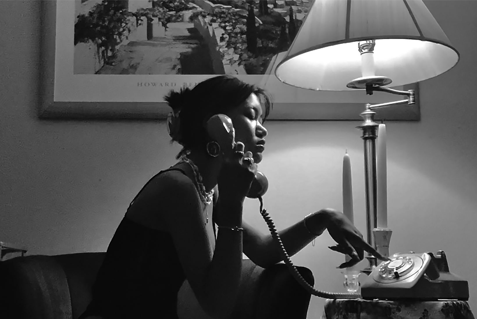 A woman sits on a living-room chair next to a table with a lamp and a taper candle and dials the rotary phone on the table. She is wearing a sleeveless shirt, and her hair is pulled back and pinned up.