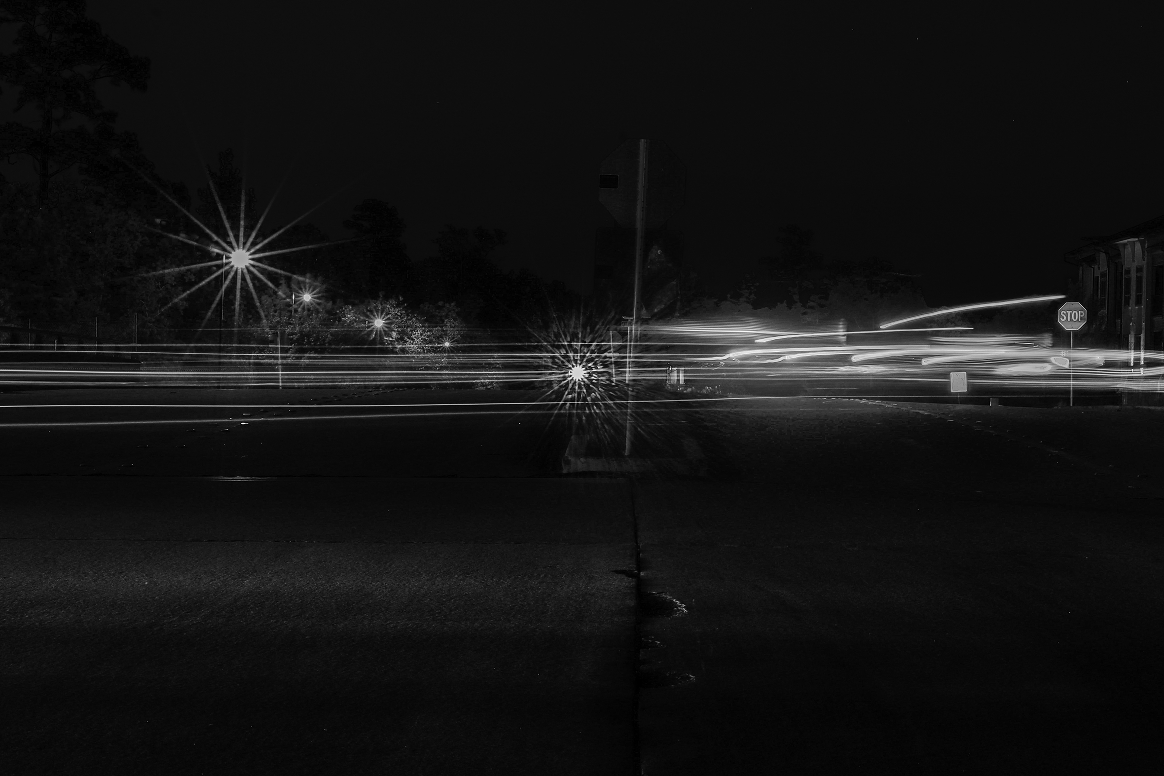 Lights shoot and zoom horizontally across a street against a dark intersection while the stationary streetlights appear as sunbursts retreating into the background.