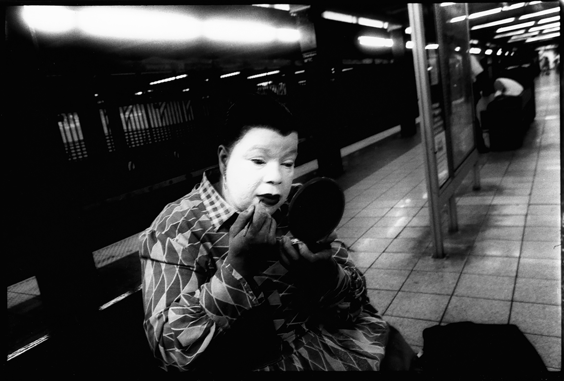 A man in white kabuki makeup sits on a subway bench and applies dark lip color with a long-handled brush as he looks into a circular handheld mirror. His glossy, black, pulled-back hair highlights a widow’s peak accentuated by the white makeup.