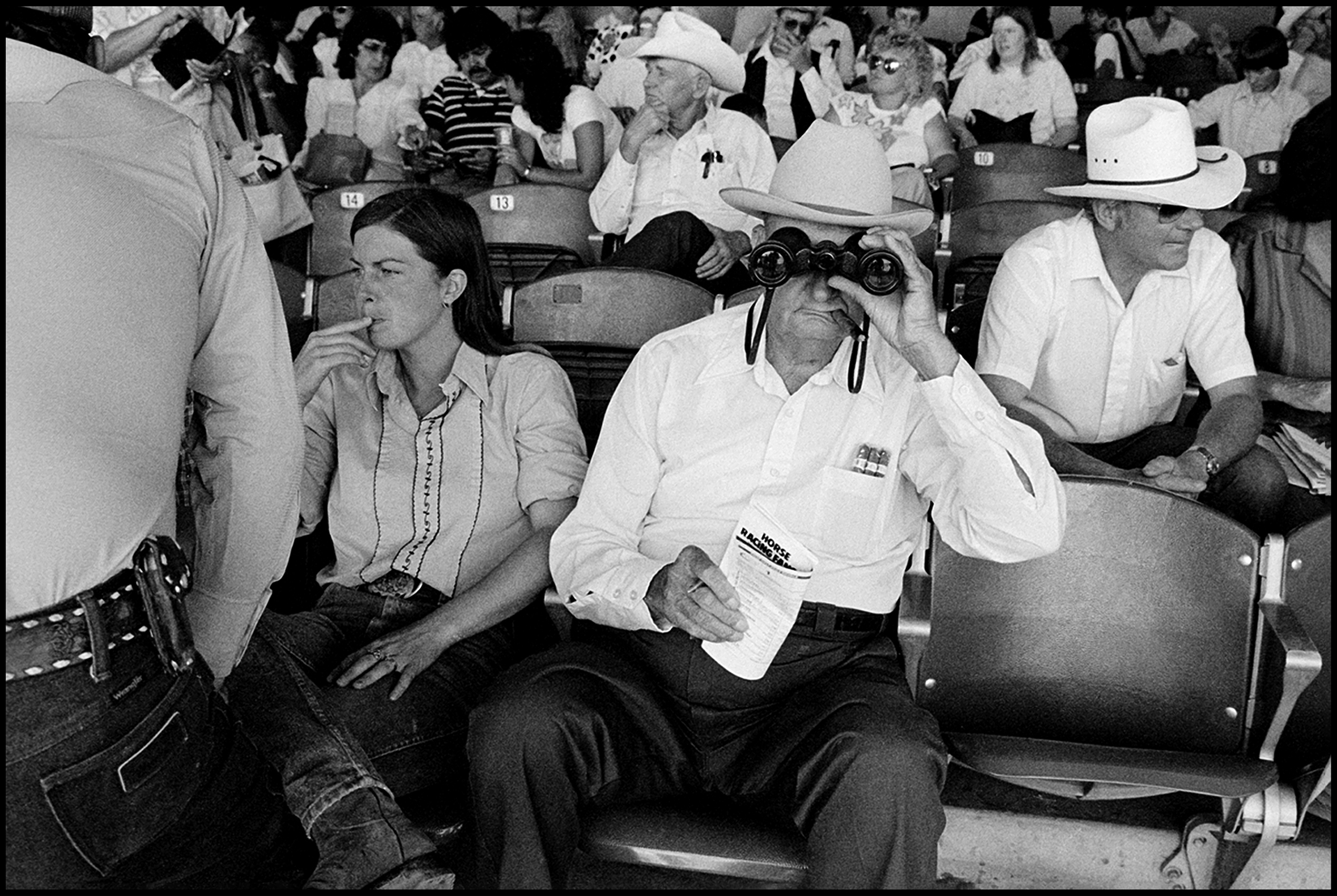 Racetrack goers sit in the stands at the Downs at Santa Fe between races. In the foreground a man and woman sit together. She looks to the side. He looks ahead through binoculars. He has a cigar in his mouth and a racing form and pen in his hand.