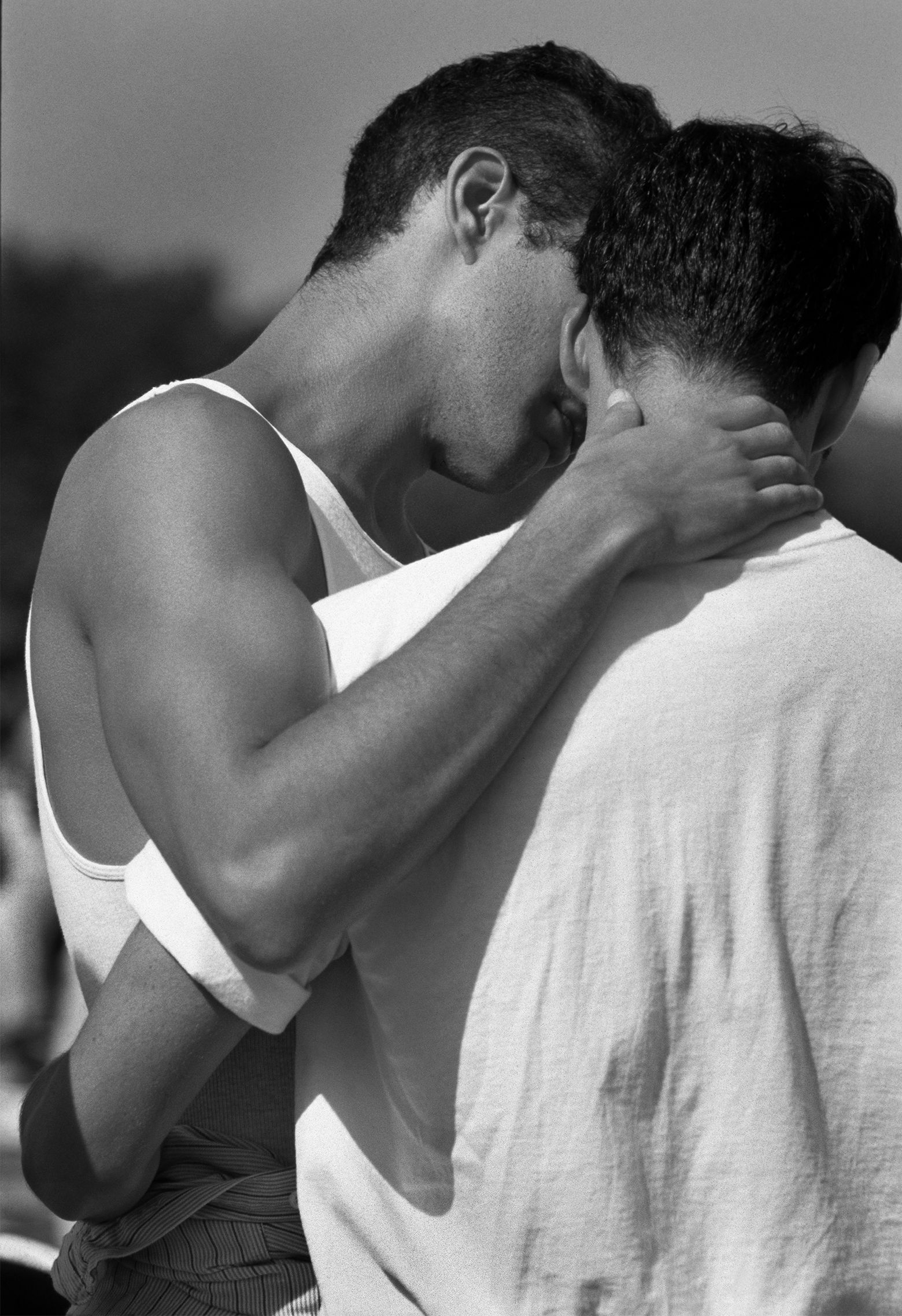 Two young men seen from the back at an angle stand closely together: one in a tank top with his hand cupped around the back of the other’s neck; one in a T-shirt with his arm around the other’s waist.